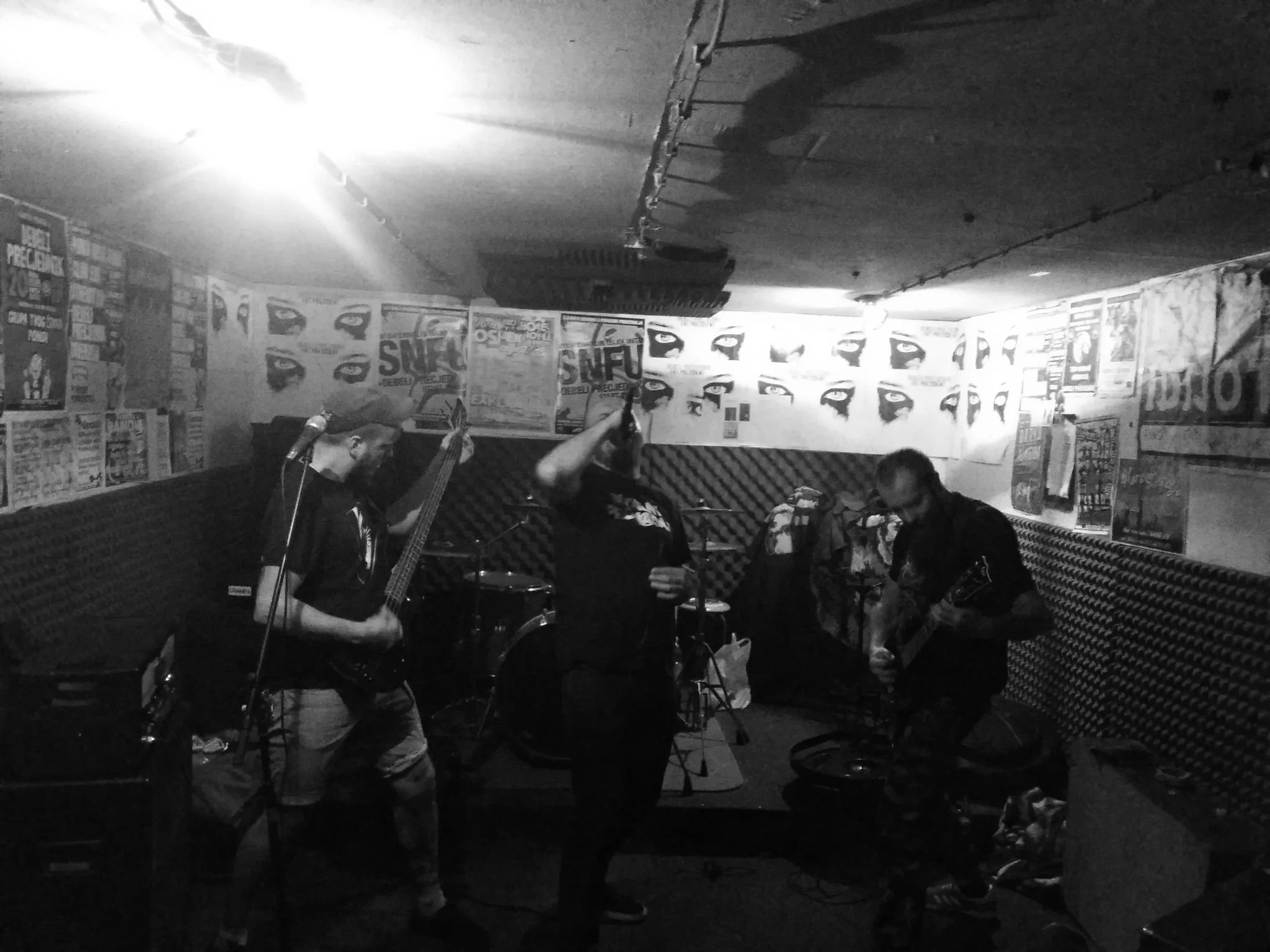 Black and white photo of band members playing instruments in a basement with posters on the walls.
