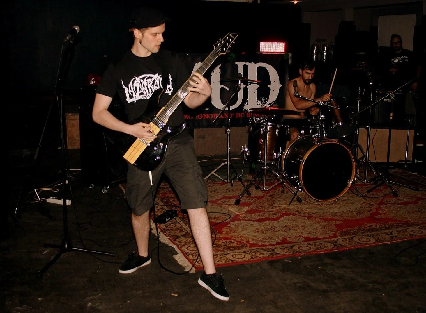 A band performing on stage with a guitarist in the foreground, a drummer in the background, and a person in the far background near the right side of the stage, and appears to be in a music venue or club.