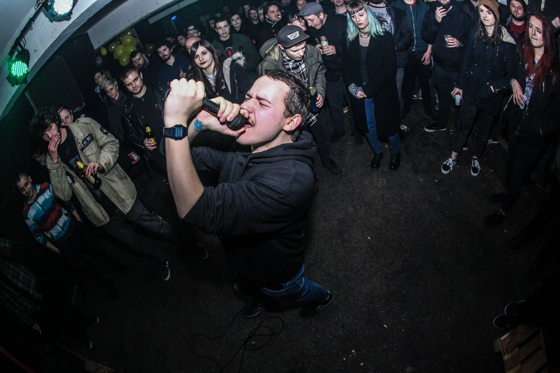 A man passionately singing into a microphone with his eyes closed, surrounded by a crowd of people at a concert or party. The crowd is watching him, some holding drinks, with diverse facial expressions. The room has low lighting with green and blue s
