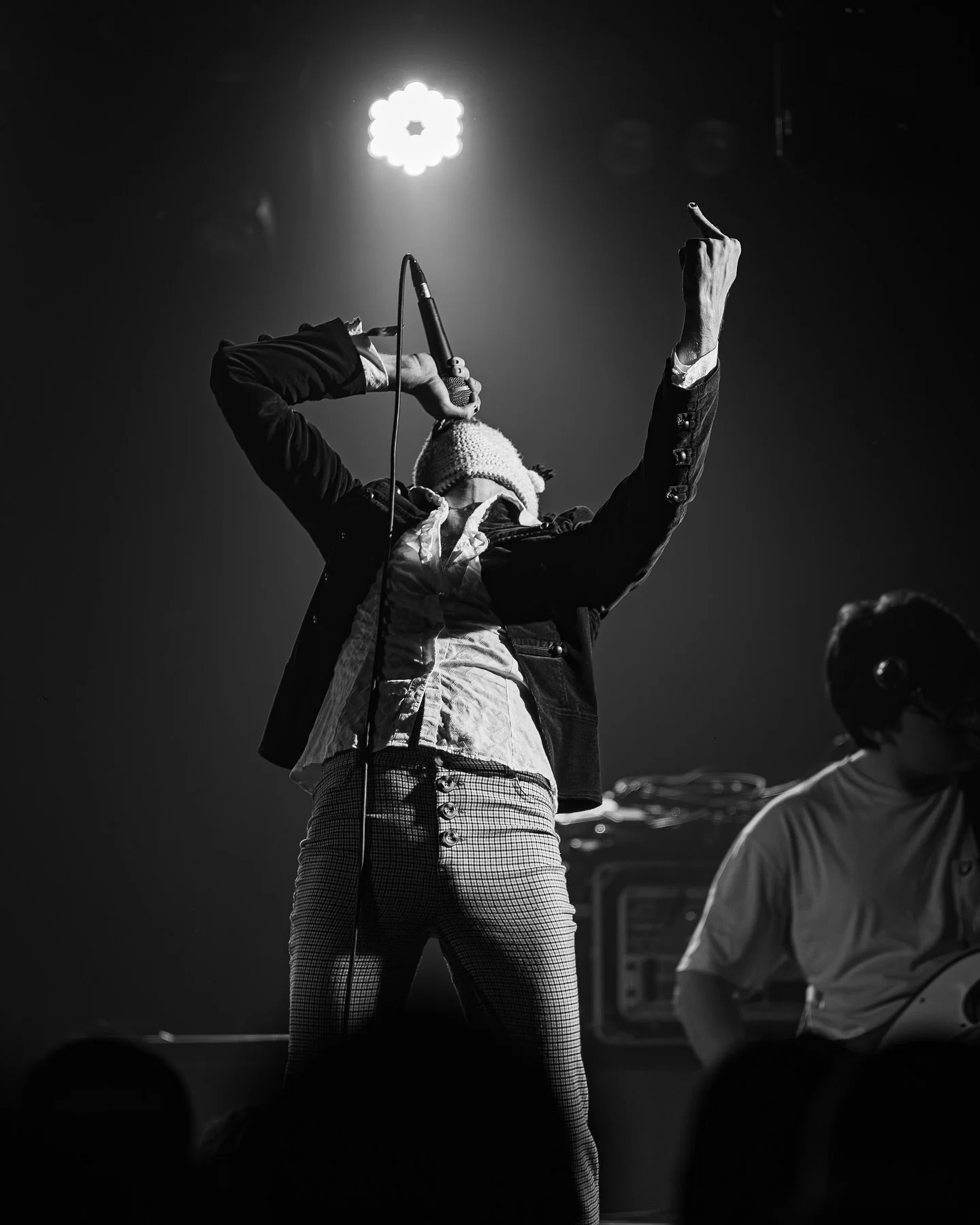 A performer on stage singing into a microphone, wearing a beanie, jacket, and plaid pants, with one arm raised and the stage lit from above in black and white.