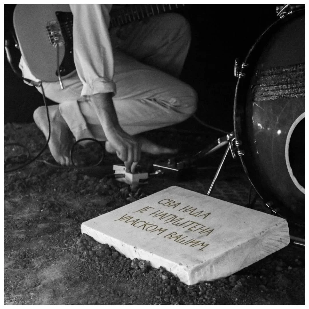 A person appears to be installing a memorial stone with inscription in Cyrillic script, next to a drum set and musical equipment at night or in a dark setting.