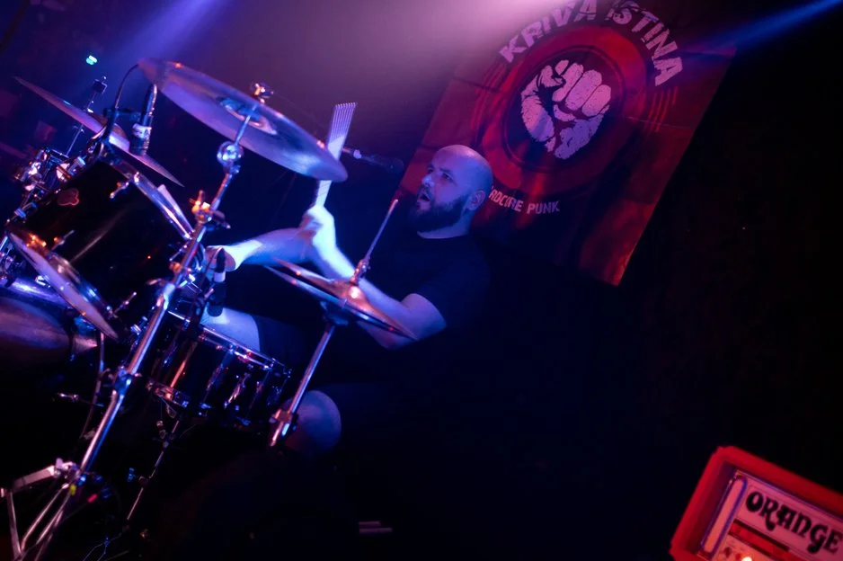 A drummer playing a drum set on stage with a red and black banner in the background that reads 'Kriva Istina' and features a clenched fist logo, illuminated by colorful stage lighting.
