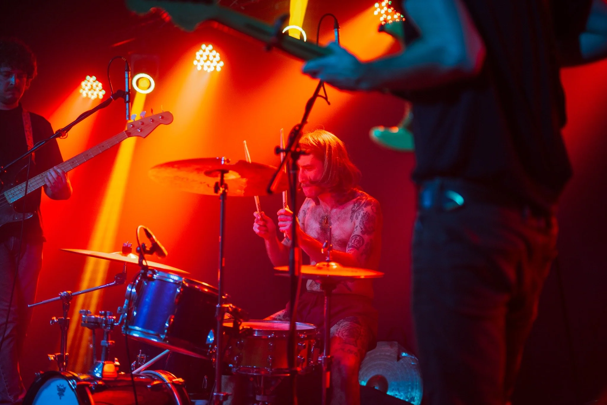 A band performing live on stage with red and orange lighting, featuring a drummer with tattoos, a guitarist, and a partially visible person in the foreground holding a string instrument.