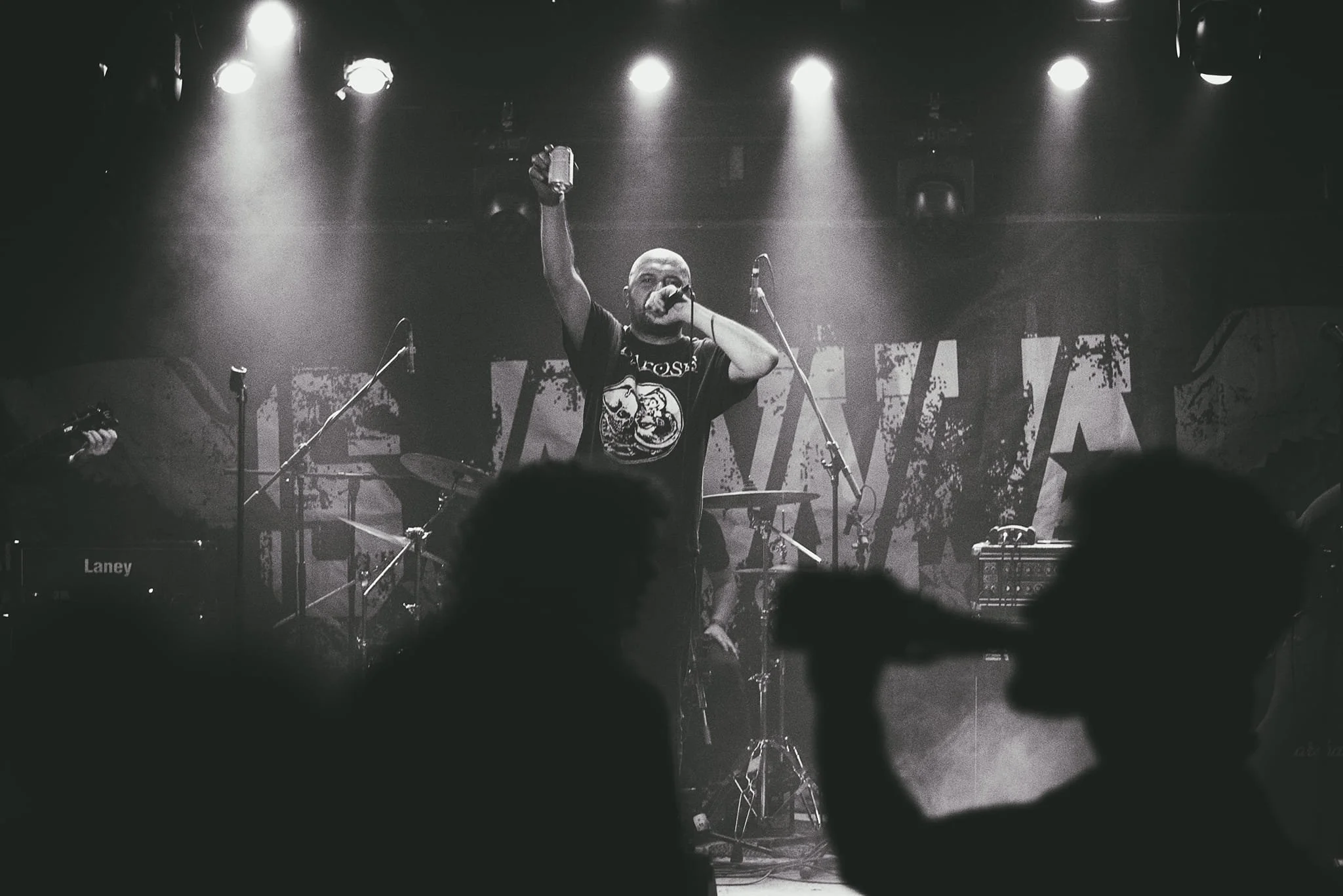 A man performs on stage at a concert, with one arm raised holding a drink and the other arm holding a microphone, surrounded by musical equipment and bright spotlights.