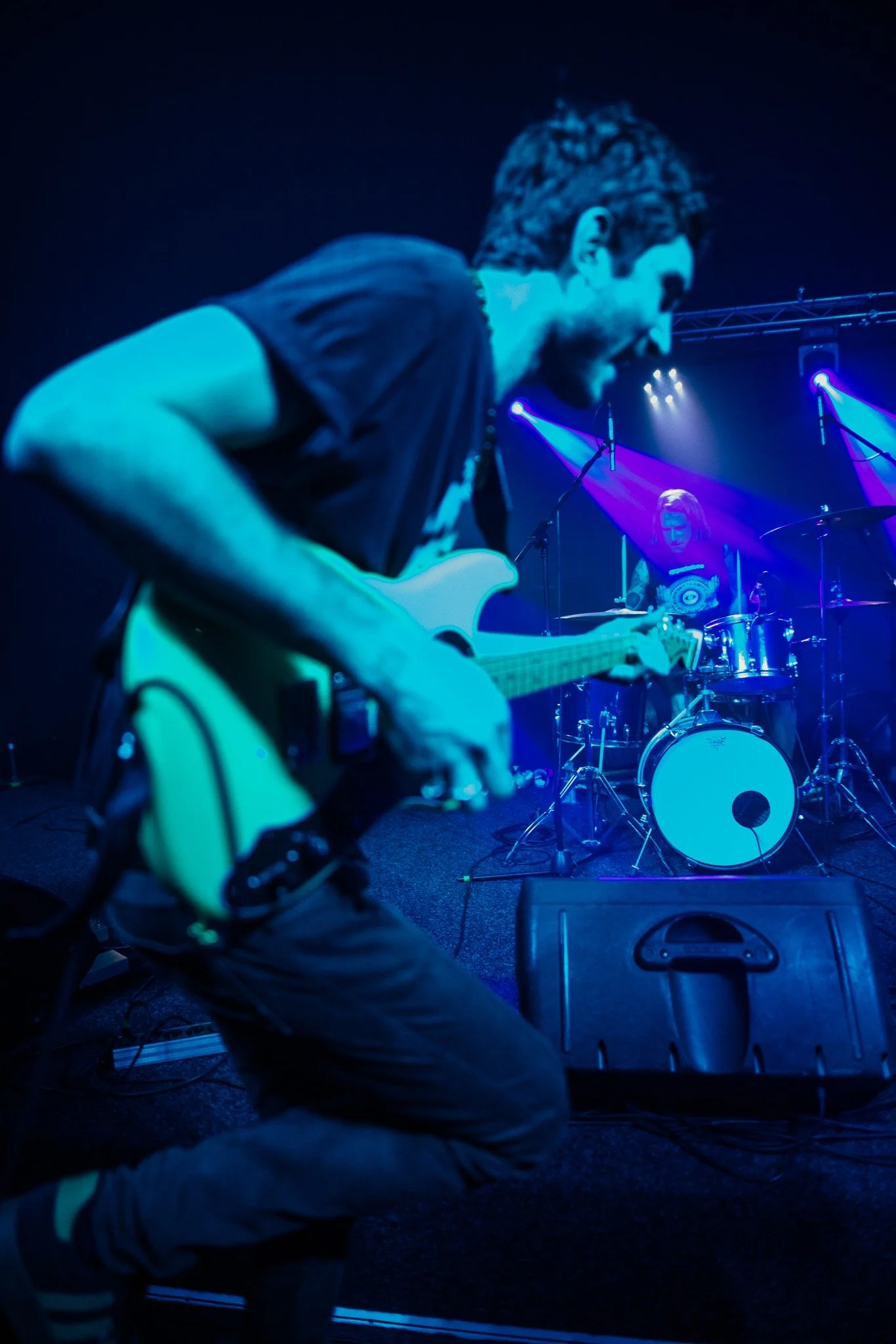 Musicians performing on stage with blue and purple lighting, one playing an electric guitar in the foreground and another on drums in the background.