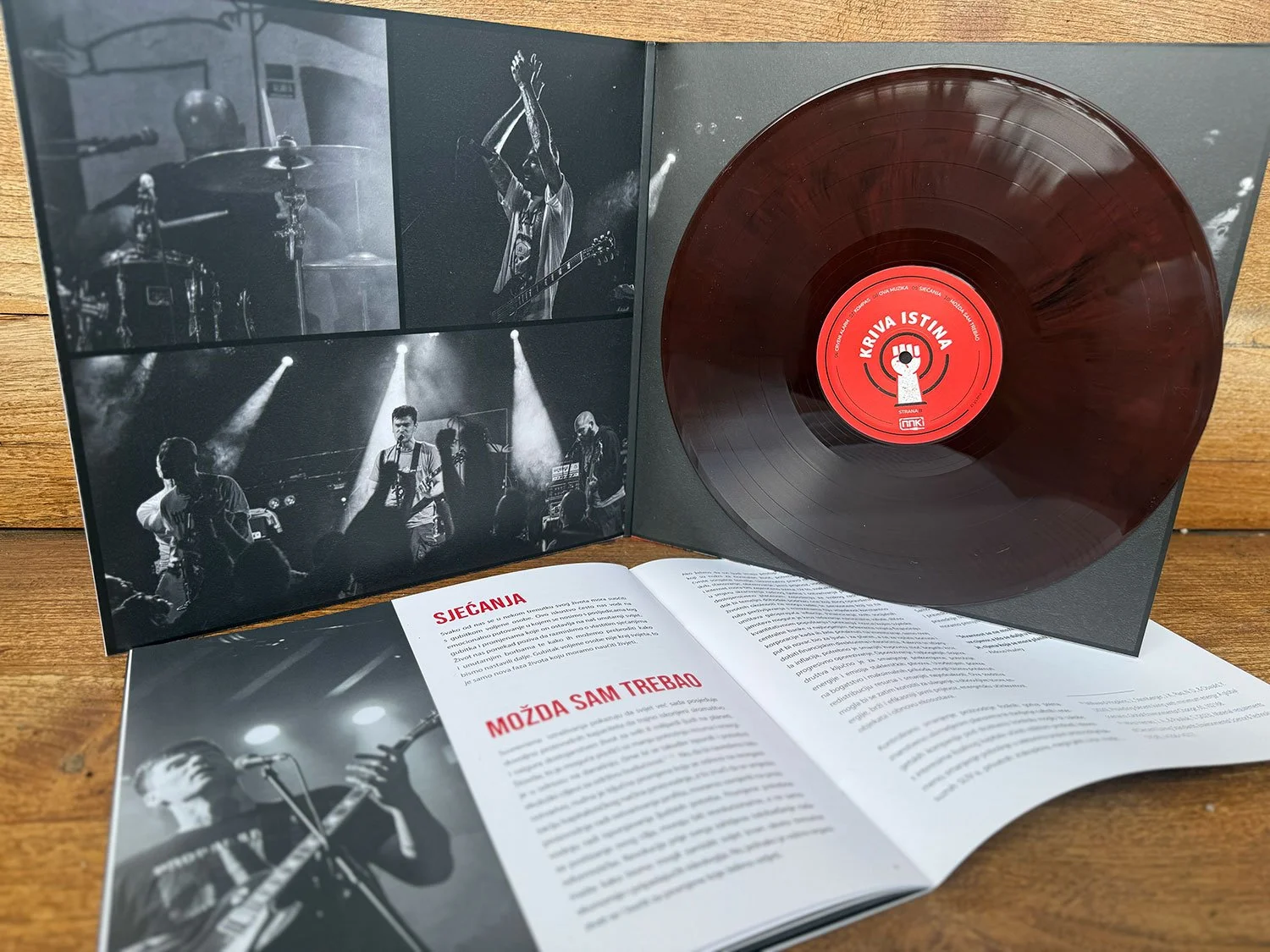 A vinyl record with a red label in a black cover, opened to show black-and-white photos of a live music performance and song lyrics in a foreign language, placed on a wooden surface.