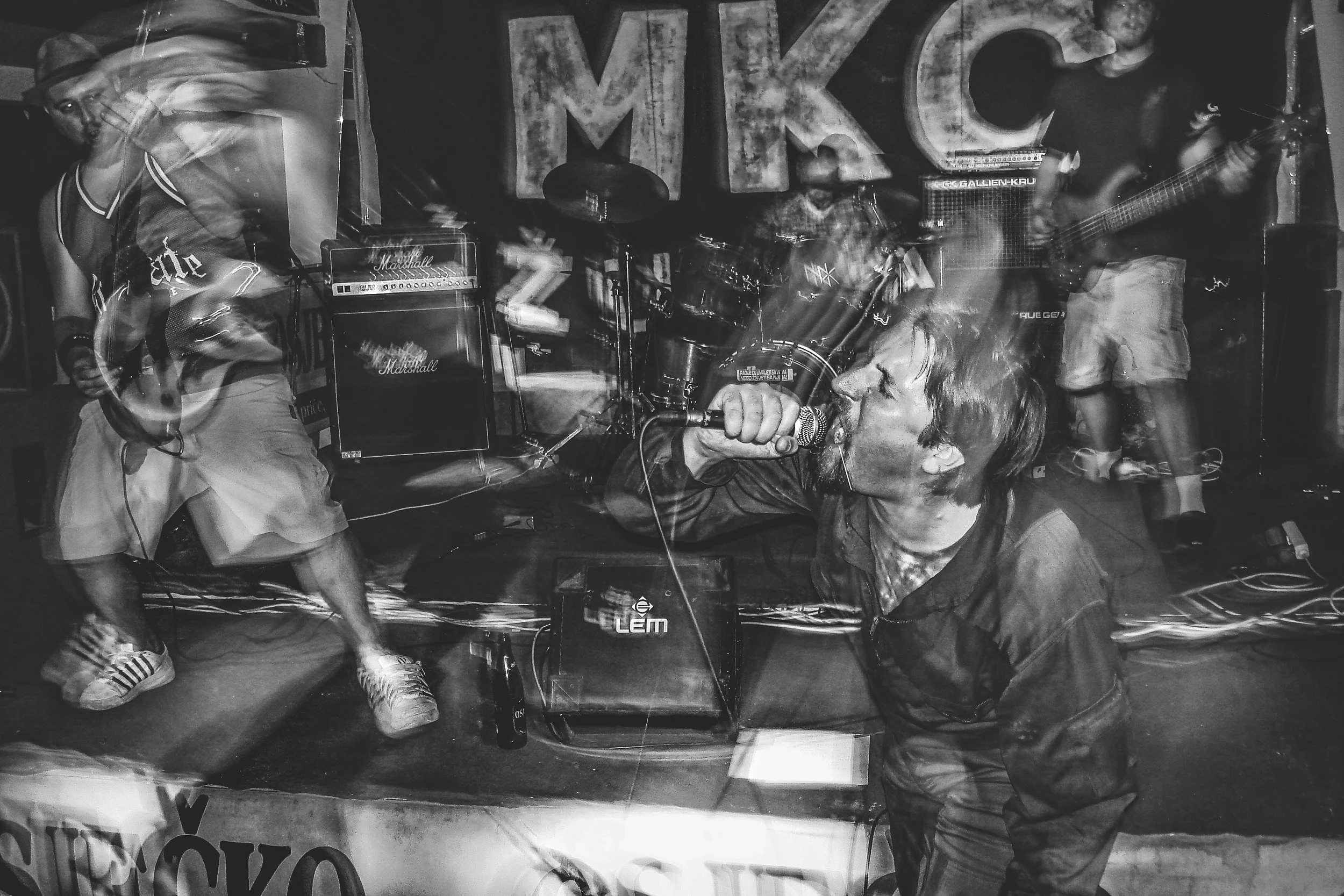 A black and white photo of a band performing on stage, featuring a vocalist with a microphone, a guitarist, and a drummer, with Amplifiers and a large sign spelling 'MIK' in the background.