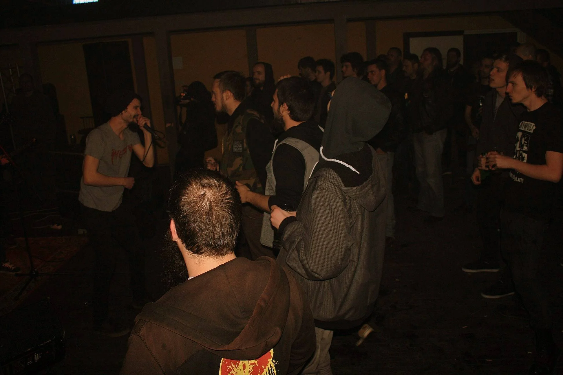 A group of people in a dimly lit indoor space, some standing and facing a person speaking into a microphone, while others are watching and listening.