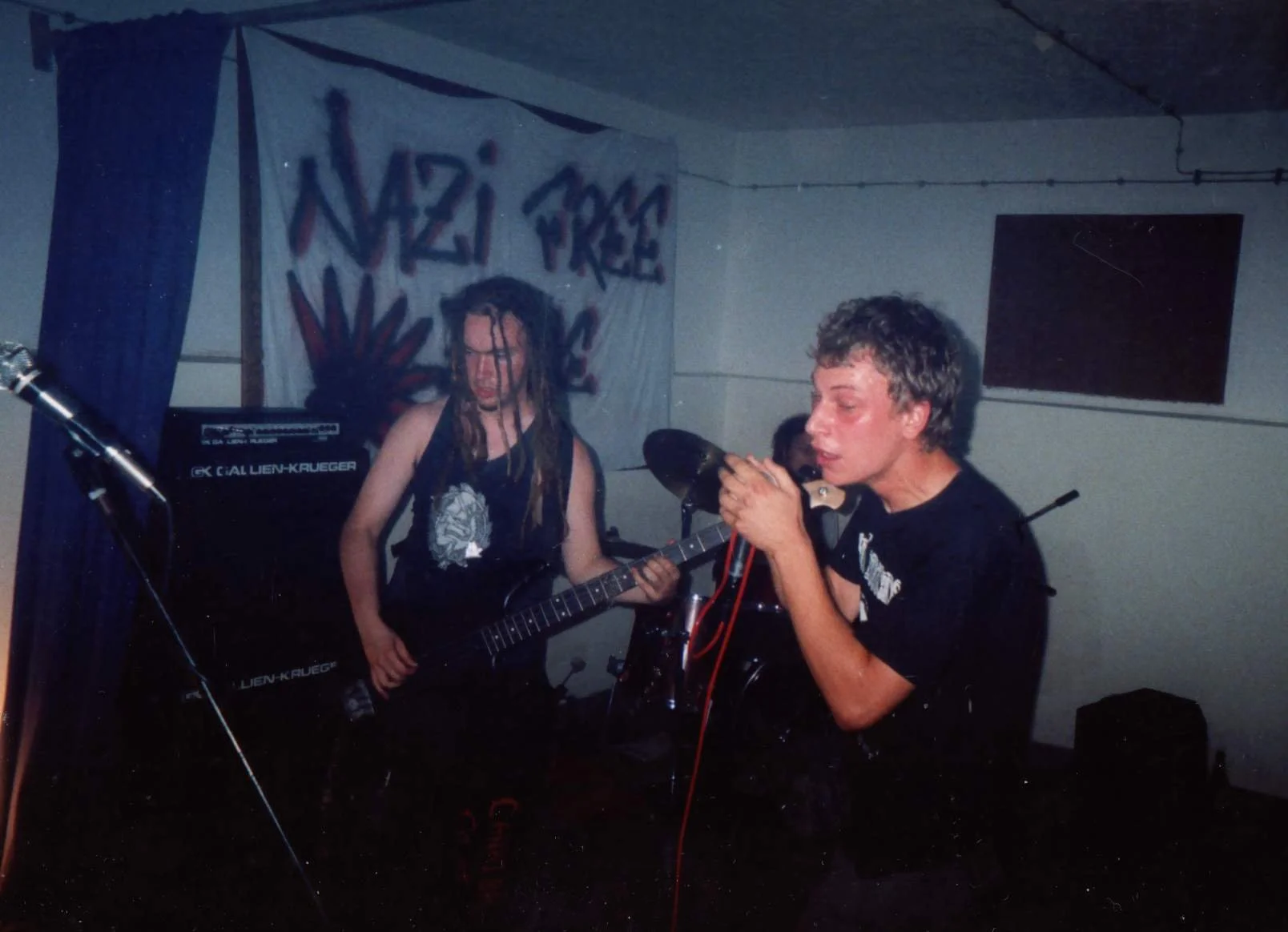 Two young men performing in a band. One is playing a guitar, the other is singing into a microphone. The background has a banner with the words 'Nazi Free' and a marijuana leaf. The setting appears to be a small, indoor venue.