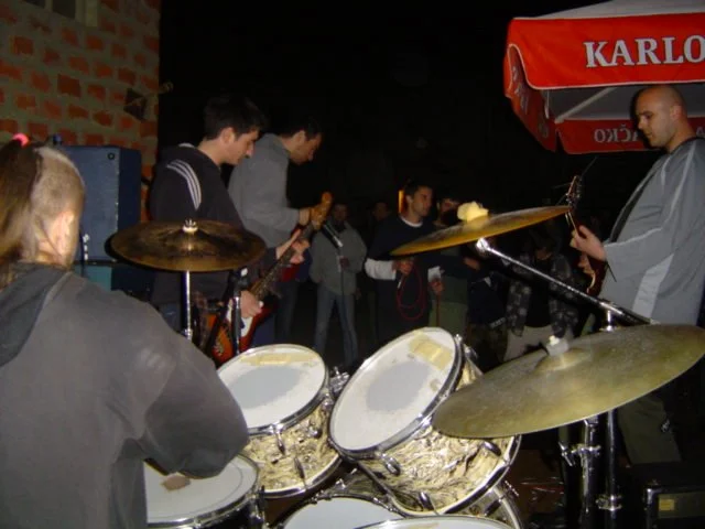 A group of people performing live music outdoors at night, with drums and a guitarist under a red canopy.