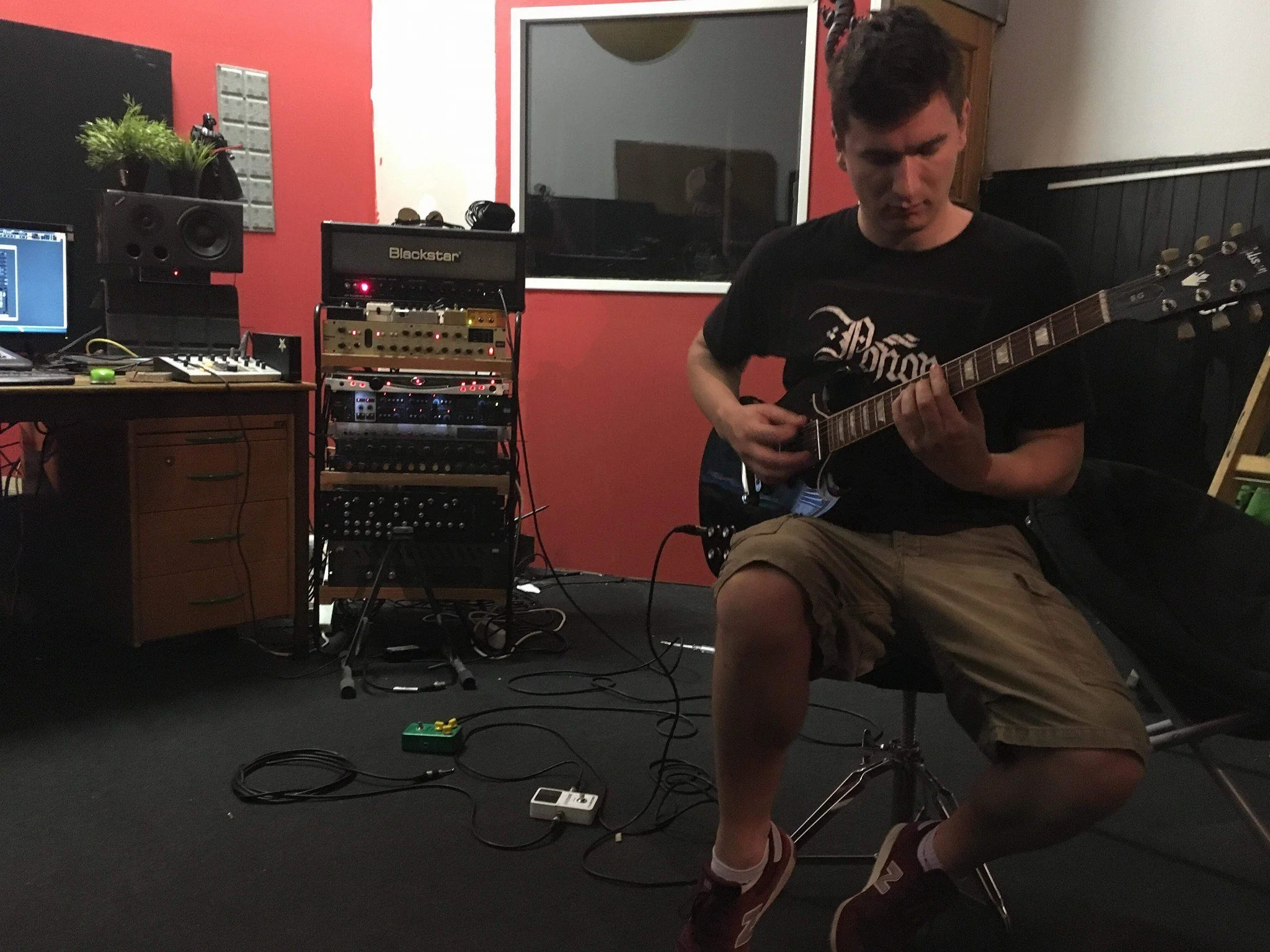 A young man with dark hair wearing a black T-shirt and tan shorts playing an electric guitar in a music studio. The studio has audio equipment, a computer, speakers, and cables, with a red and white wall in the background.
