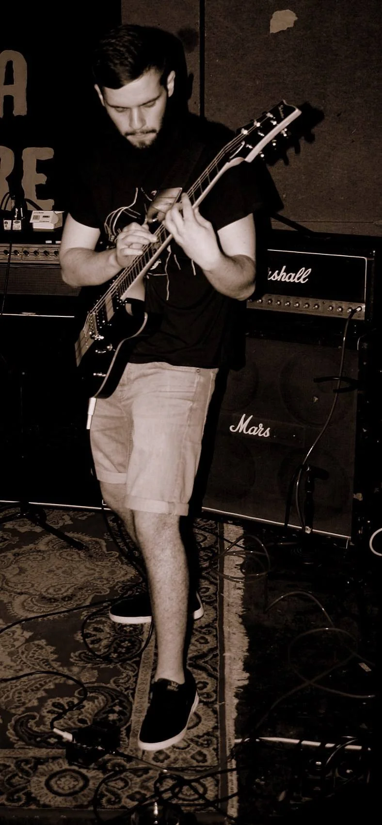 A young man playing an electric guitar on stage, with Marshall amplifiers in the background, wearing a black t-shirt, beige shorts, and black sneakers.