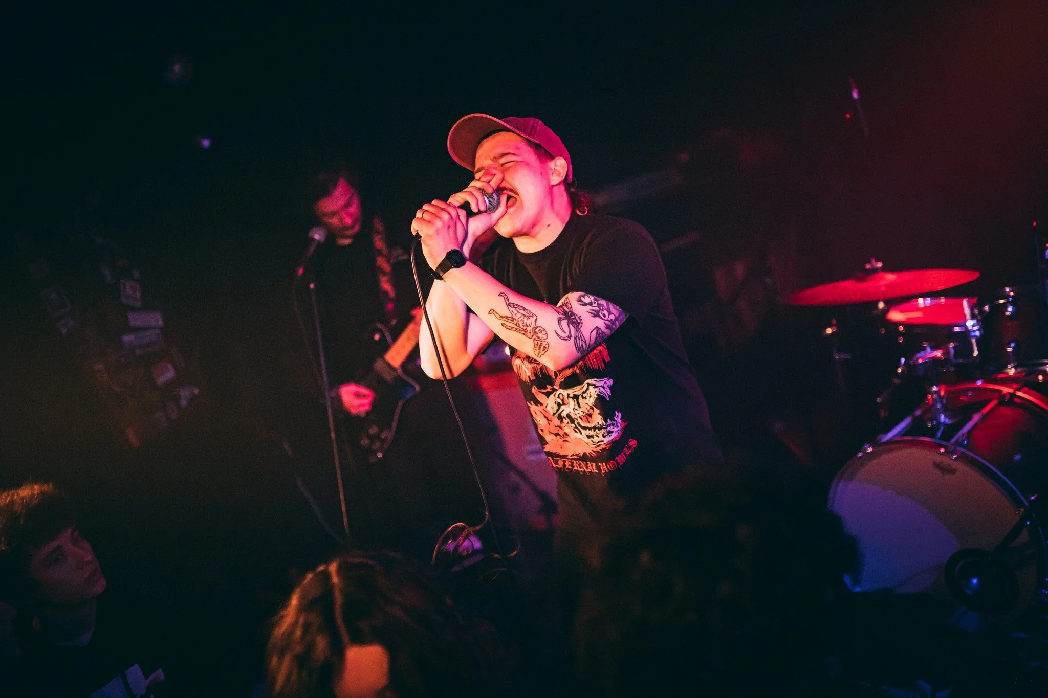 A person passionately singing into a microphone during a live music performance at a dimly lit venue, with a drum set visible in the background and audience members watching.