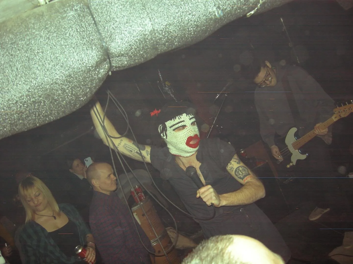 A singer with a mask singing into a microphone on a stage with a guitarist and an audience in a dark venue.