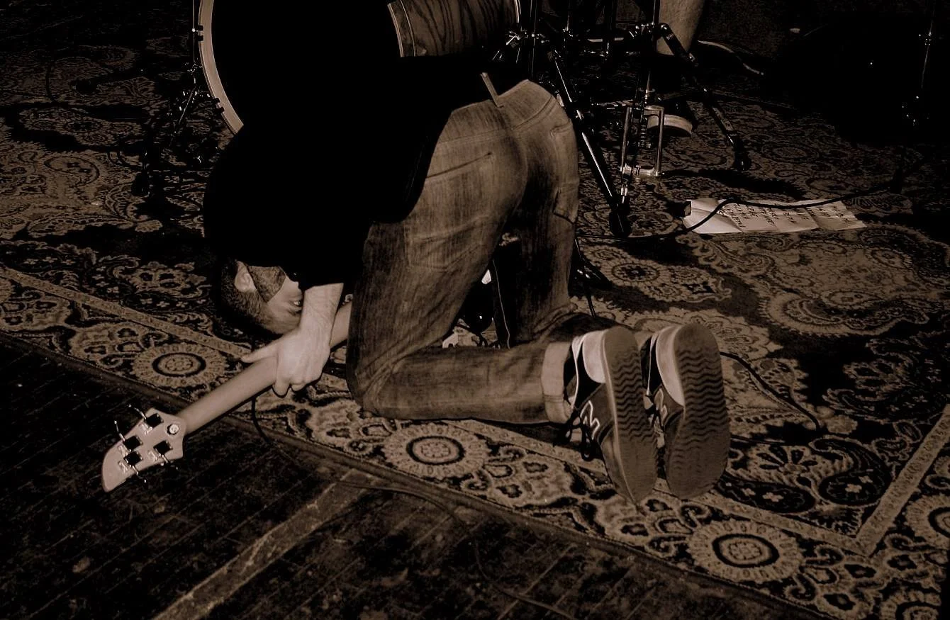 A person kneeling on a patterned rug, clutching an electric guitar, with a drum set in the background at a music venue or recording studio.