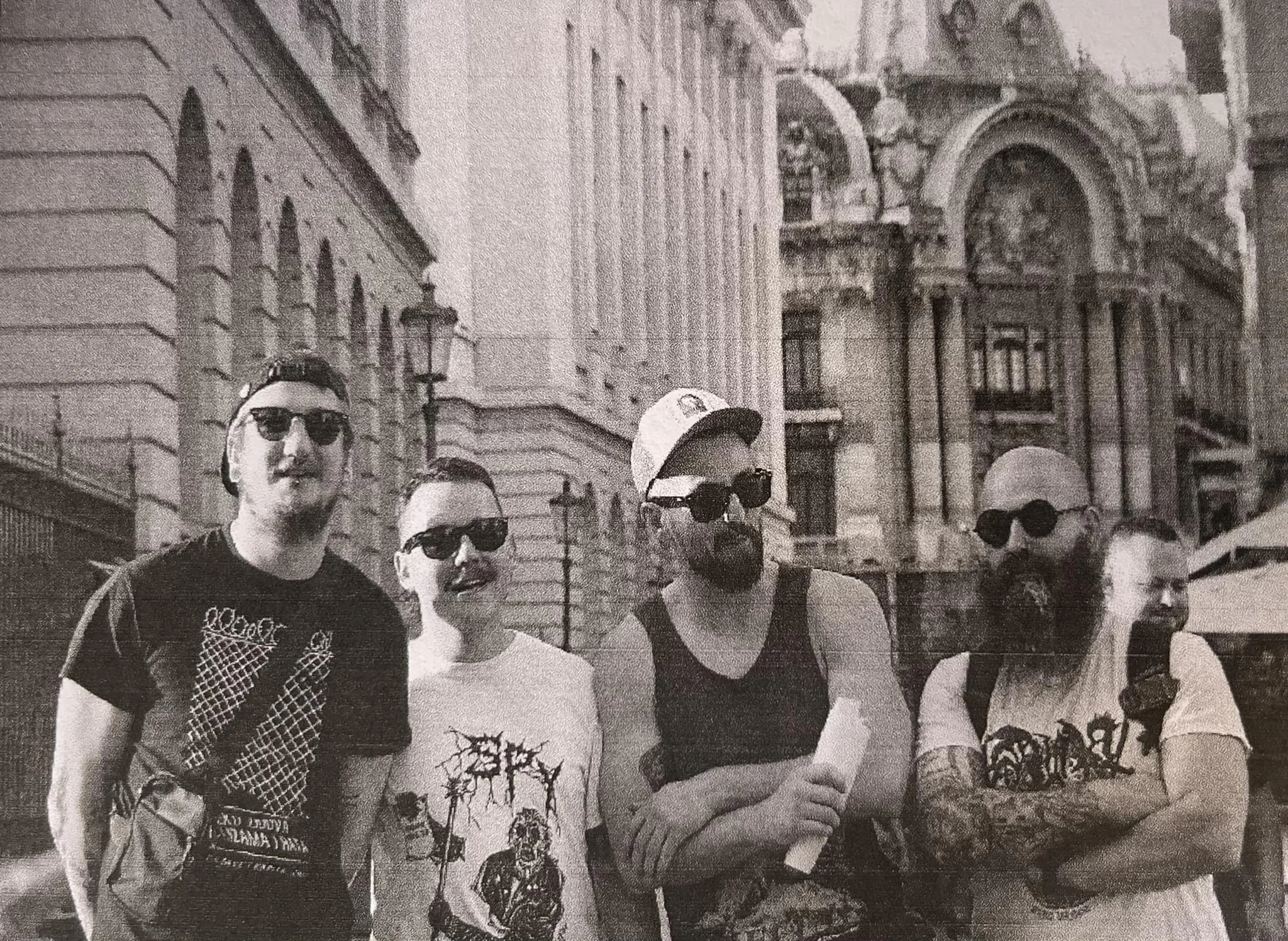 A group of young men standing outdoors on a city street, wearing sunglasses and casual clothing, with historic buildings in the background.