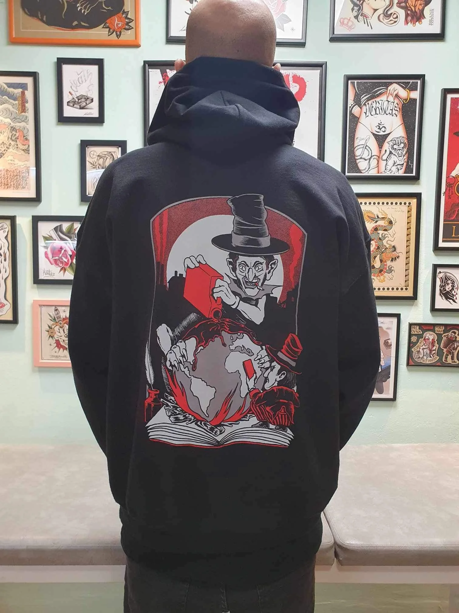A person with a shaved head wearing a hoodie with a graphic of a magician pulling the world out of a hat, standing in front of a wall with framed artwork.