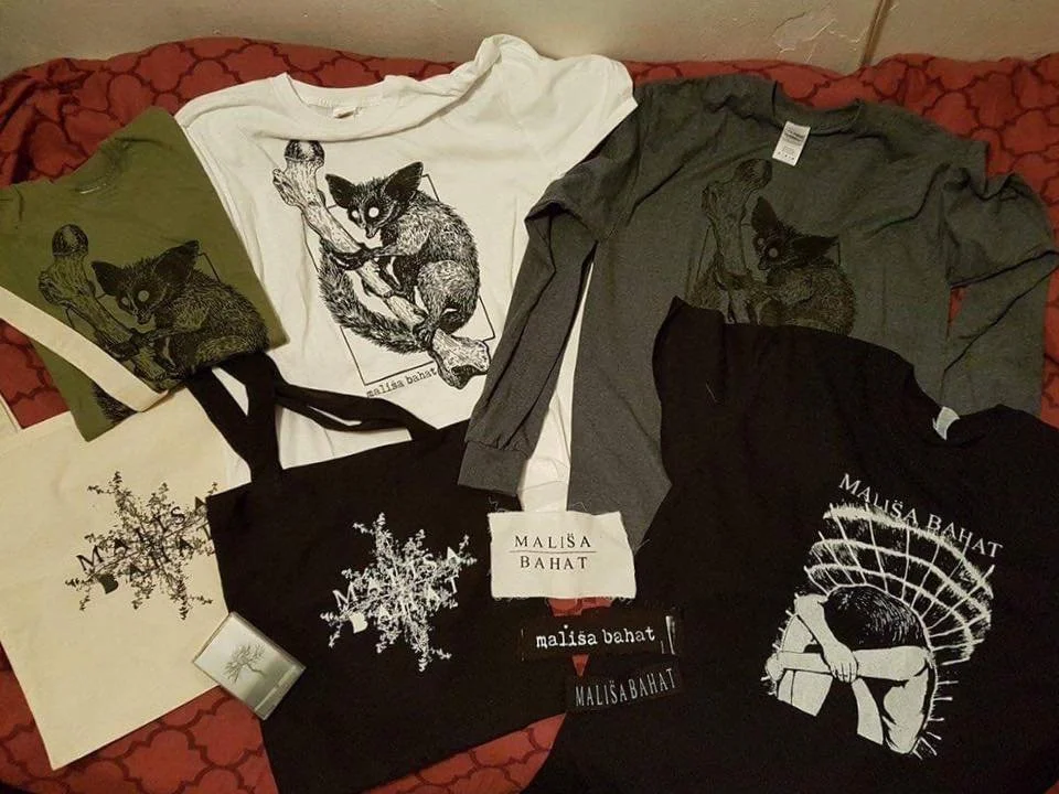 Various shirts and tote bags with black and white or colored prints of cats and trees, all labeled with the name 'Mališa Bahat' on a red patterned fabric surface.