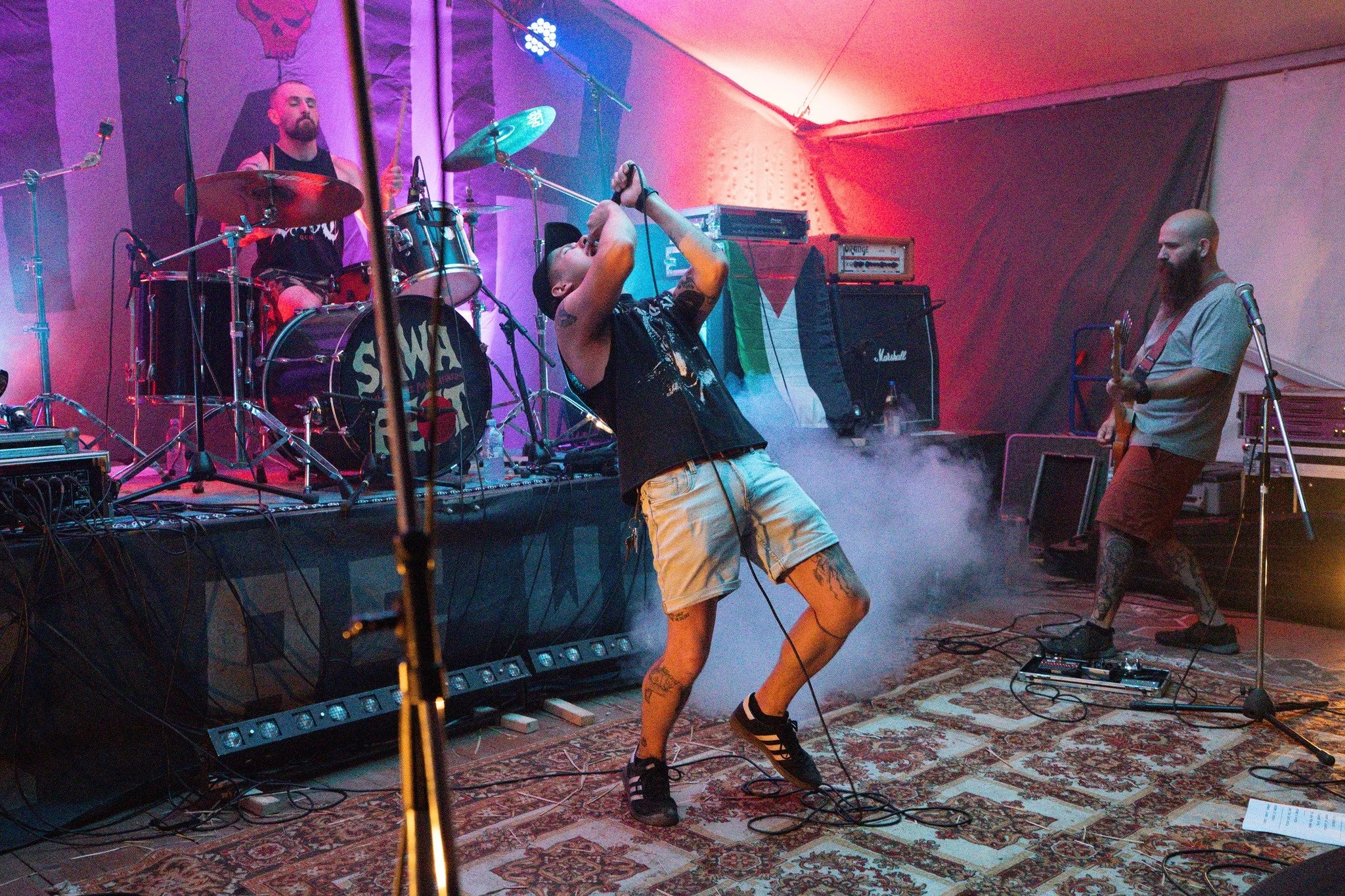 A band performing live on stage with a singer, guitarist, and drummer. The singer is shirtless, holding a microphone, and appears to be singing passionately. The drummer is seated behind a drum kit, and the guitarist is on the right side of the stage