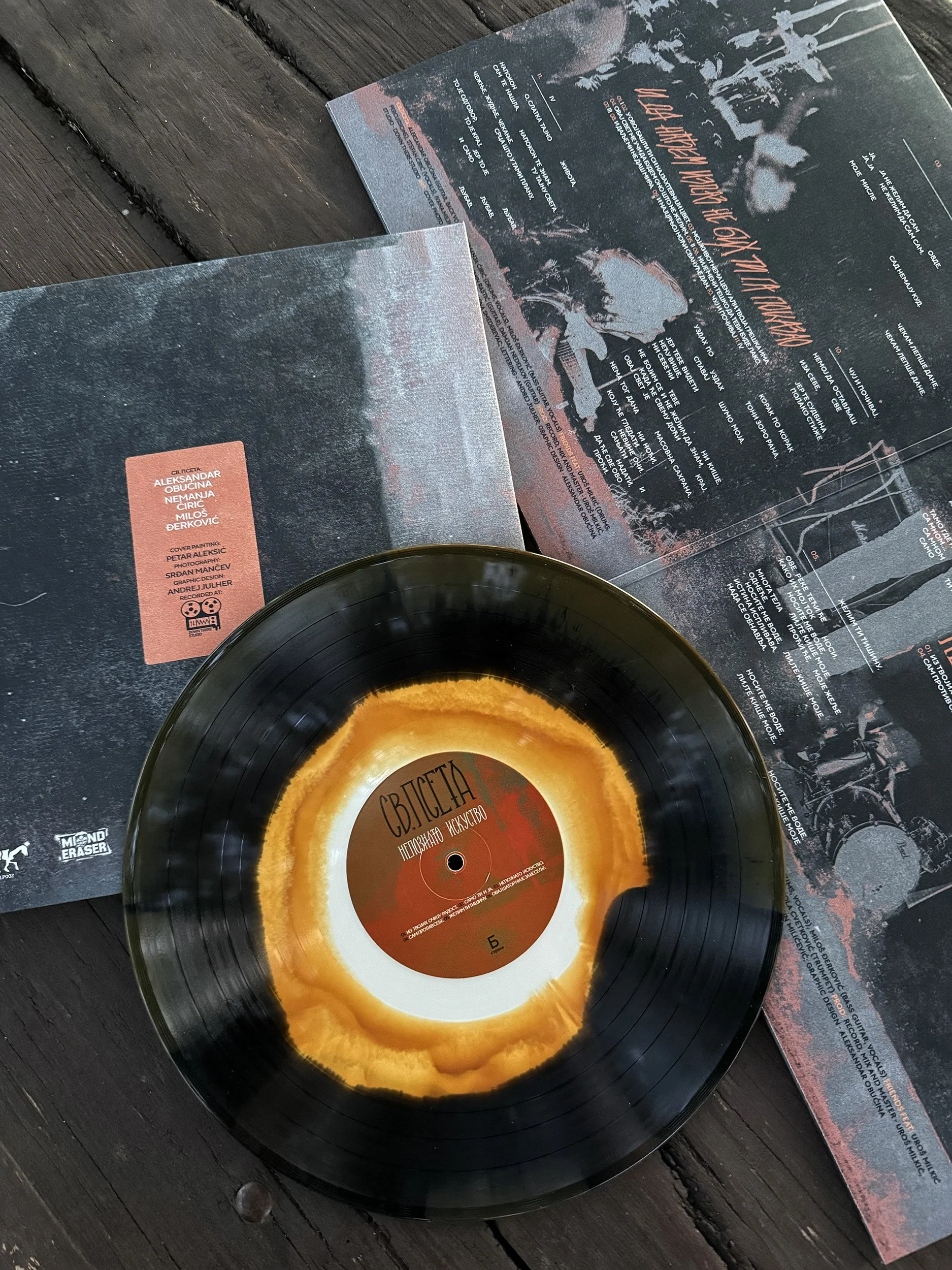 A vinyl record with a yellow and brown center label is placed on a dark wooden surface, partially covering a musical album cover or booklet with text and artwork. Band name: SV. PSETA / SVETA PSETA