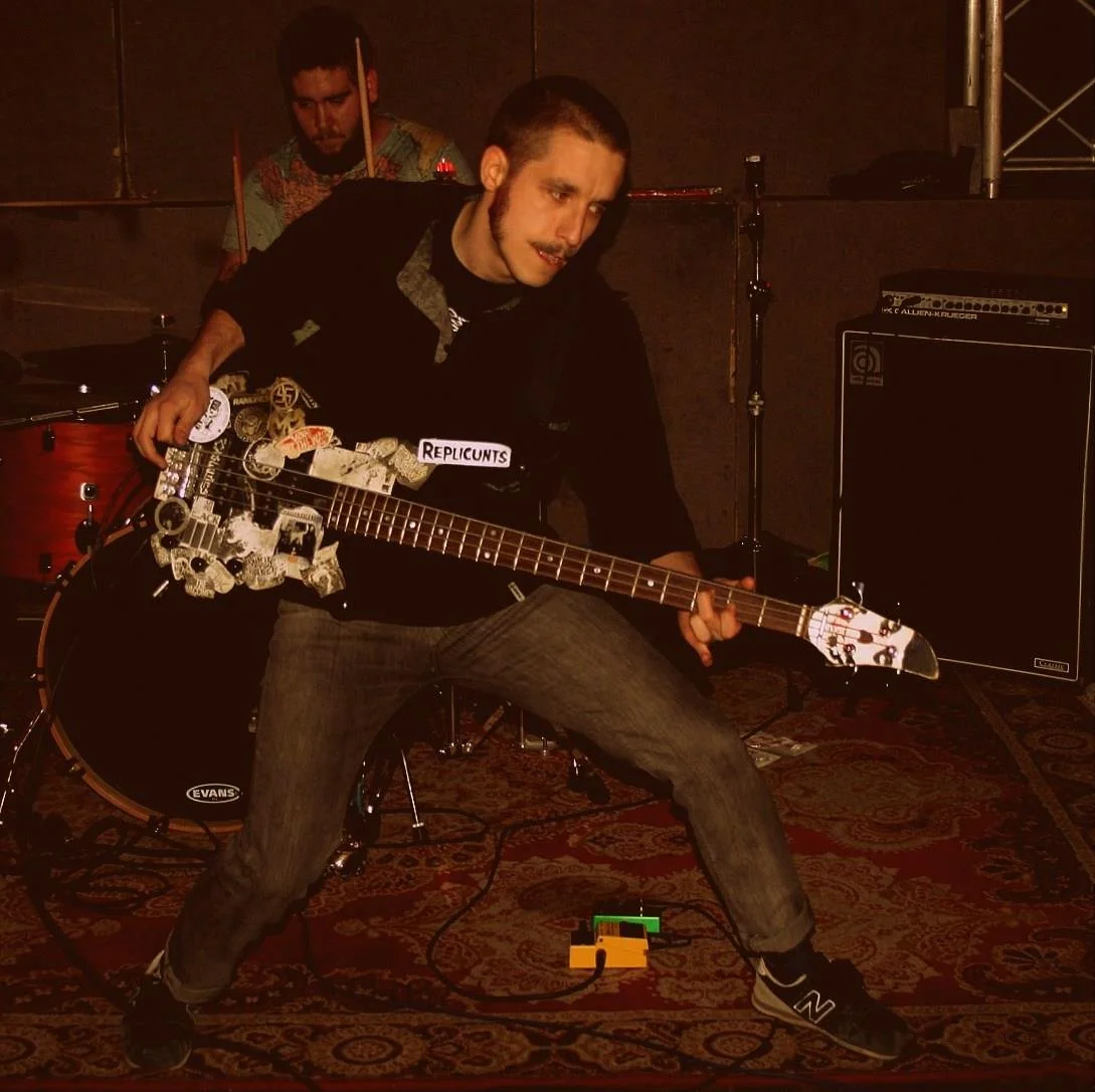 Young man playing an electric guitar with stickers, wearing a black hoodie and gray pants, in a music rehearsal space. In the background, a drummer is visible, and musical equipment such as an amplifier and a drum set are present.