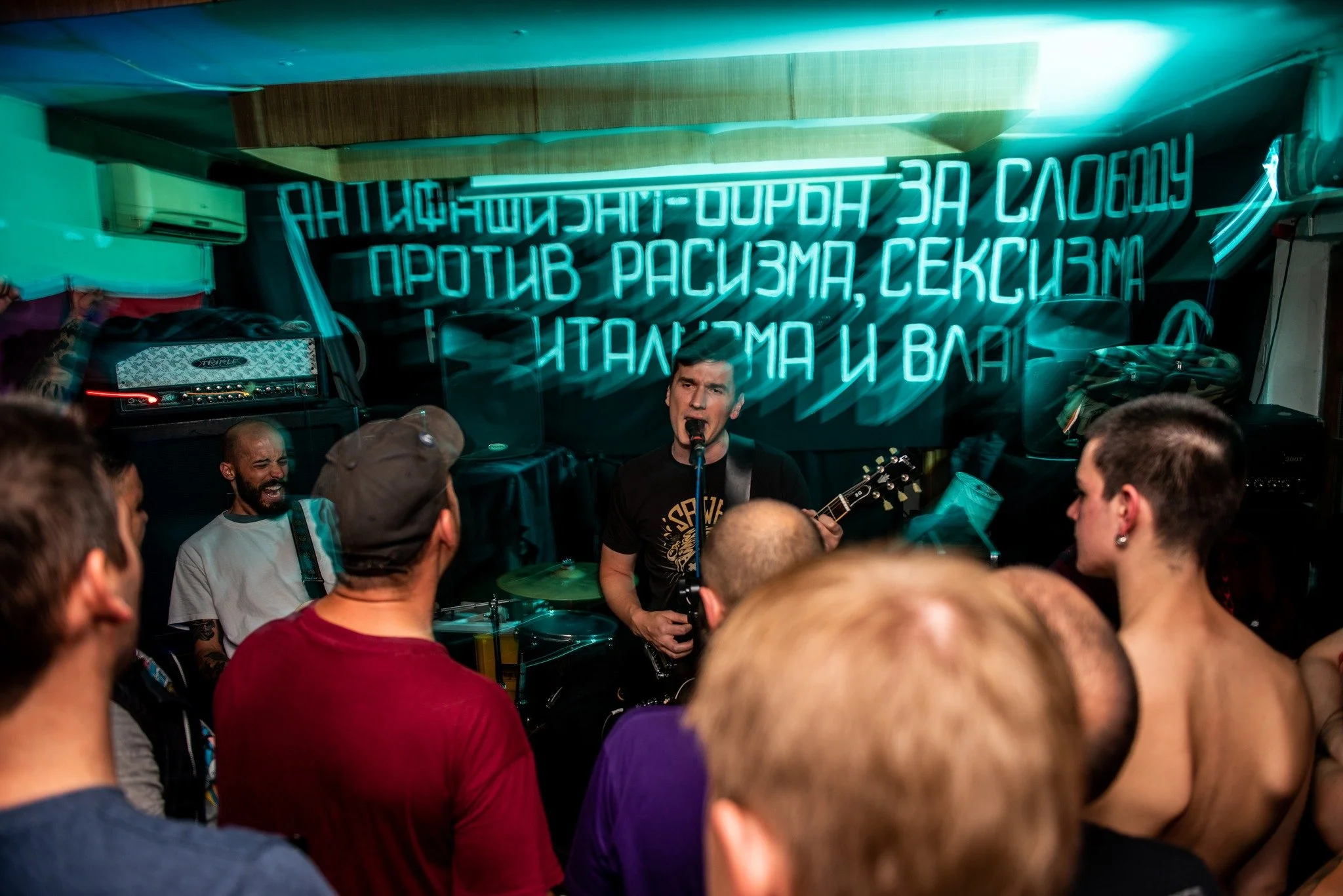 A crowded room with a band performing live music. The band includes a singer playing guitar and a drummer. There is a bright neon sign in the background with text in a foreign language.