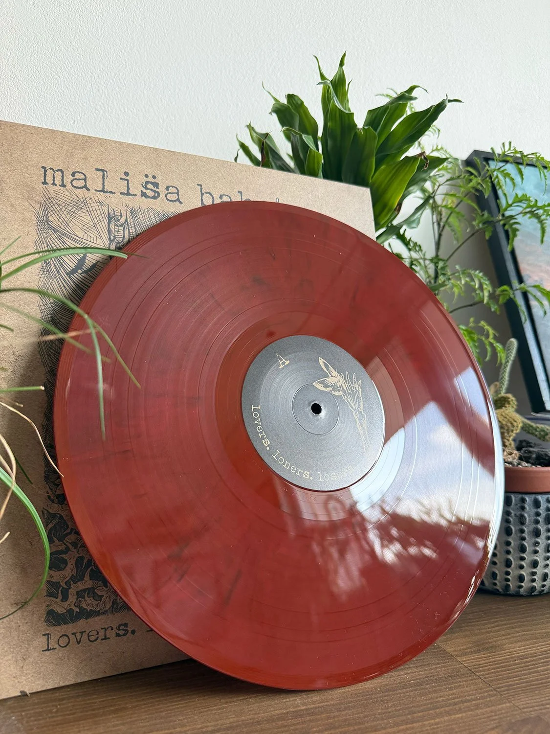 Red vinyl record on a wooden surface with a label that reads "Lovers" and an illustration of a unicorn's head, surrounded by green potted plants in the background.