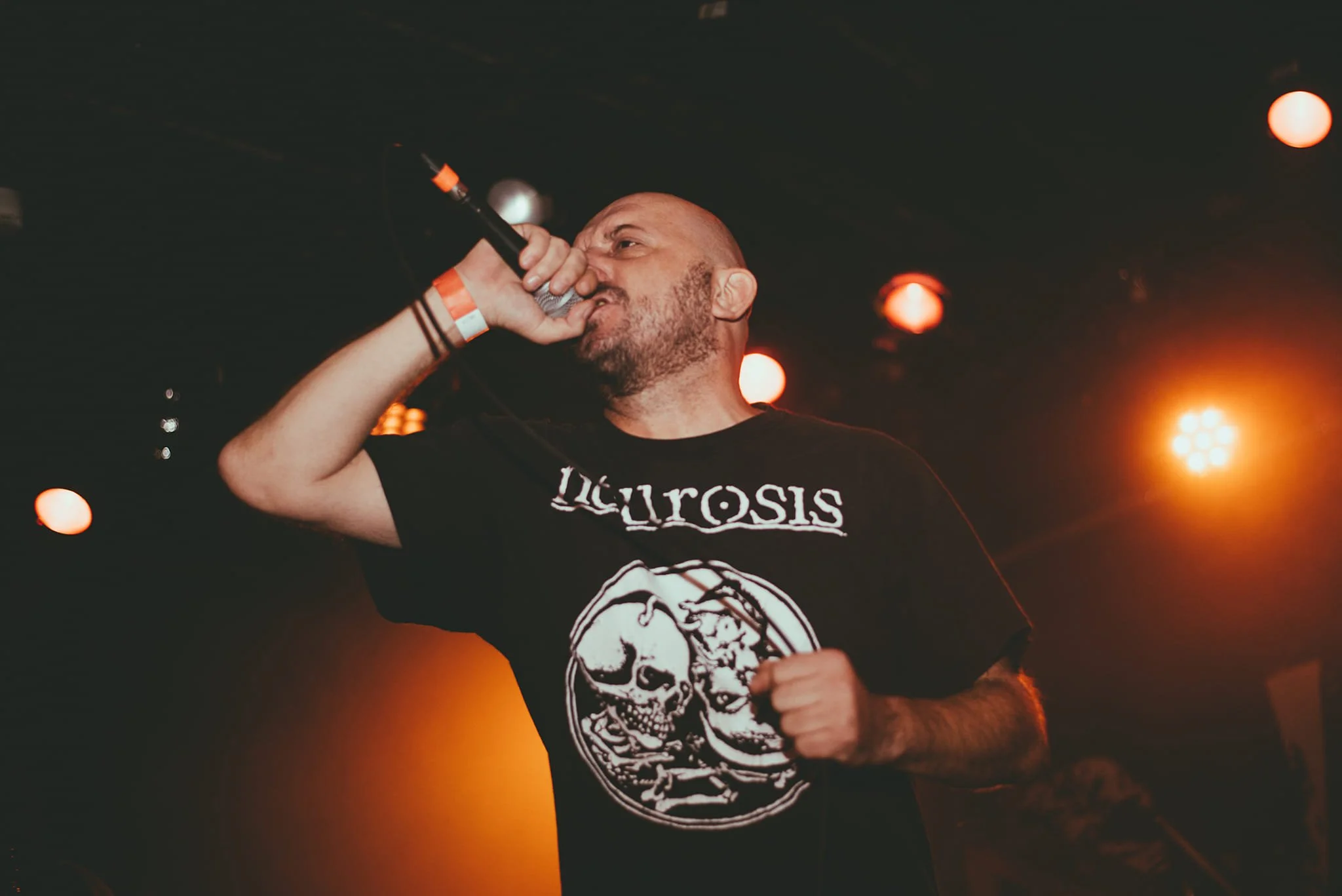 A man with a tattoo sleeve on his left arm is singing into a microphone on stage with orange stage lights illuminating the background.