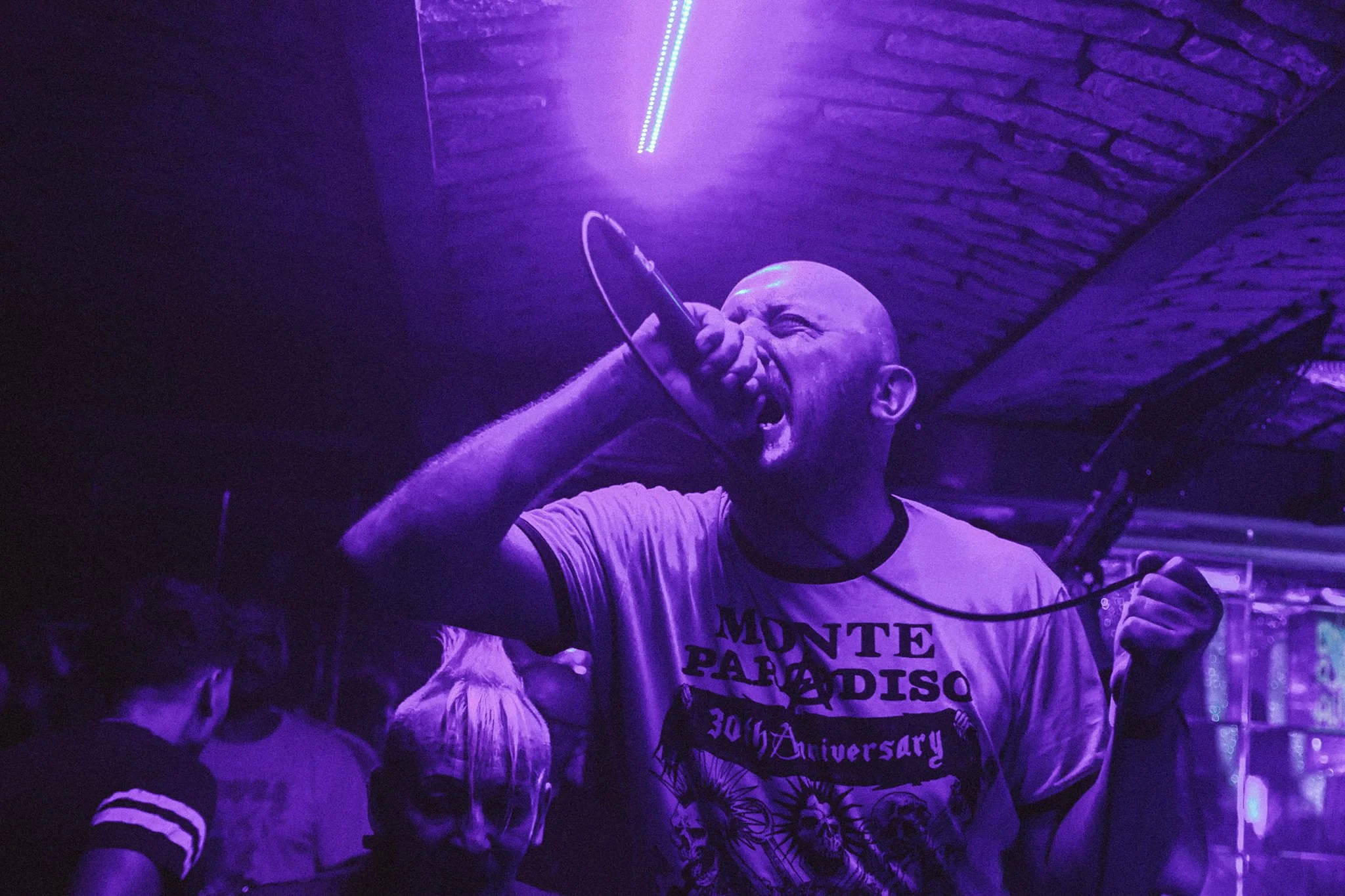 A man with a shaved head singing or shouting into a microphone in a dimly lit setting with purple lighting, wearing a shirt with the words "Monte Paradiso" and "30th Anniversary" printed on it.