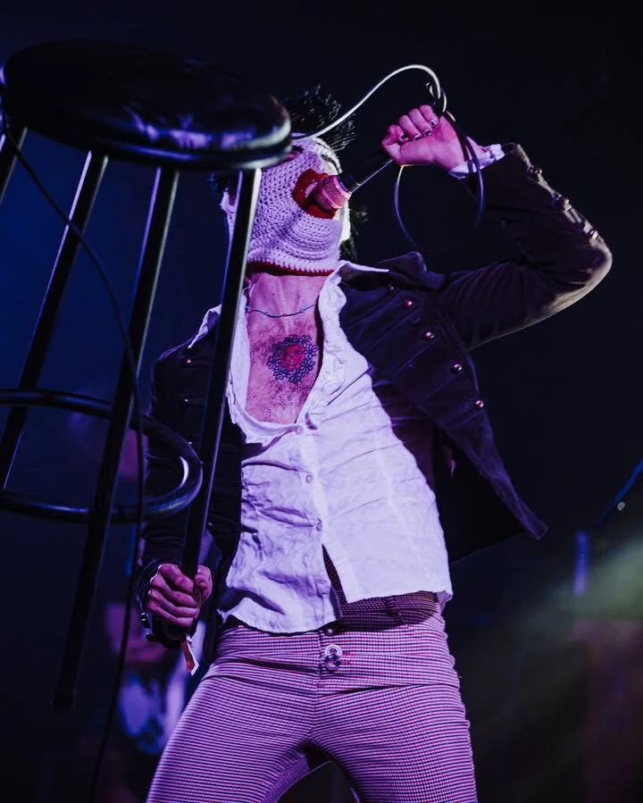Performer singing into a microphone on stage, wearing a crochet mask, black jacket, white shirt, and striped pants, with stage lighting in the background.