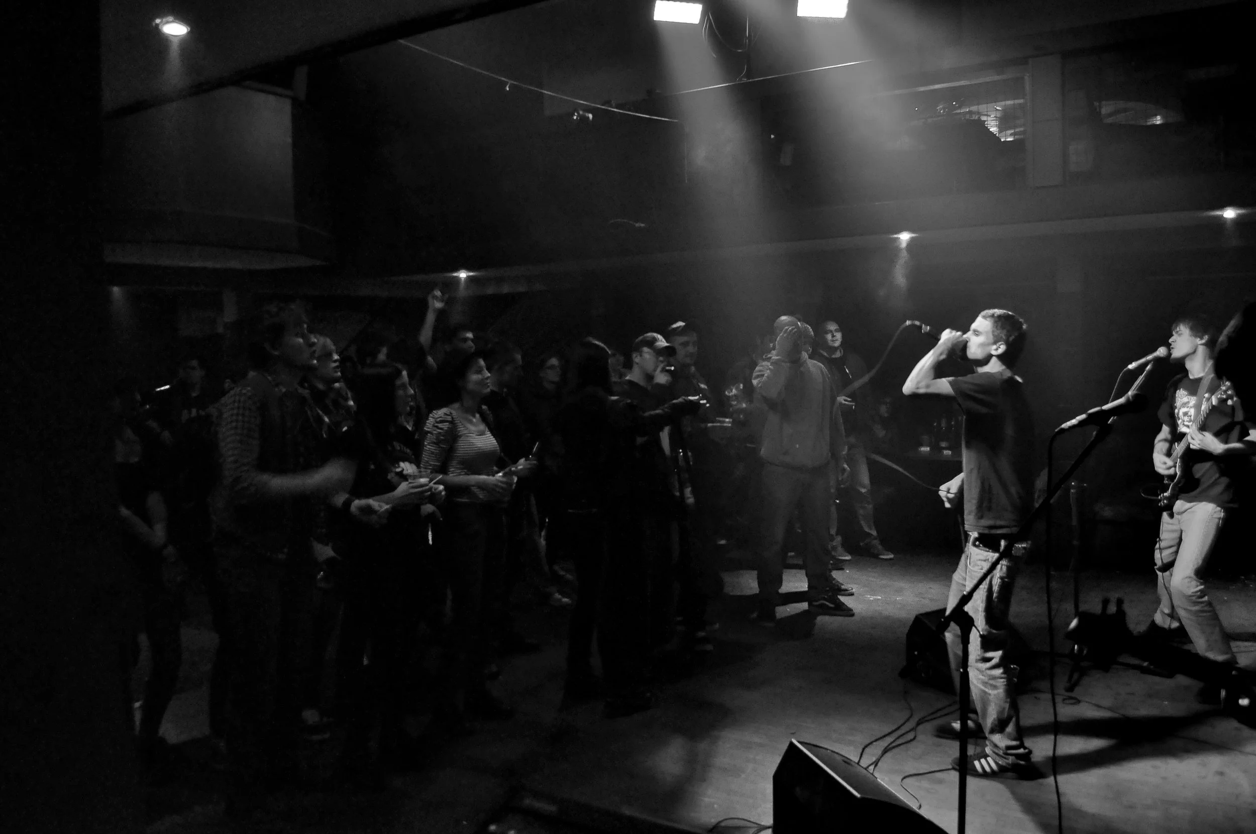 A black and white image of a small concert or performance in a dimly lit indoor venue, featuring two musicians on stage with microphones, one playing an acoustic guitar and the other singing. The audience, consisting of mostly young adults, faces the