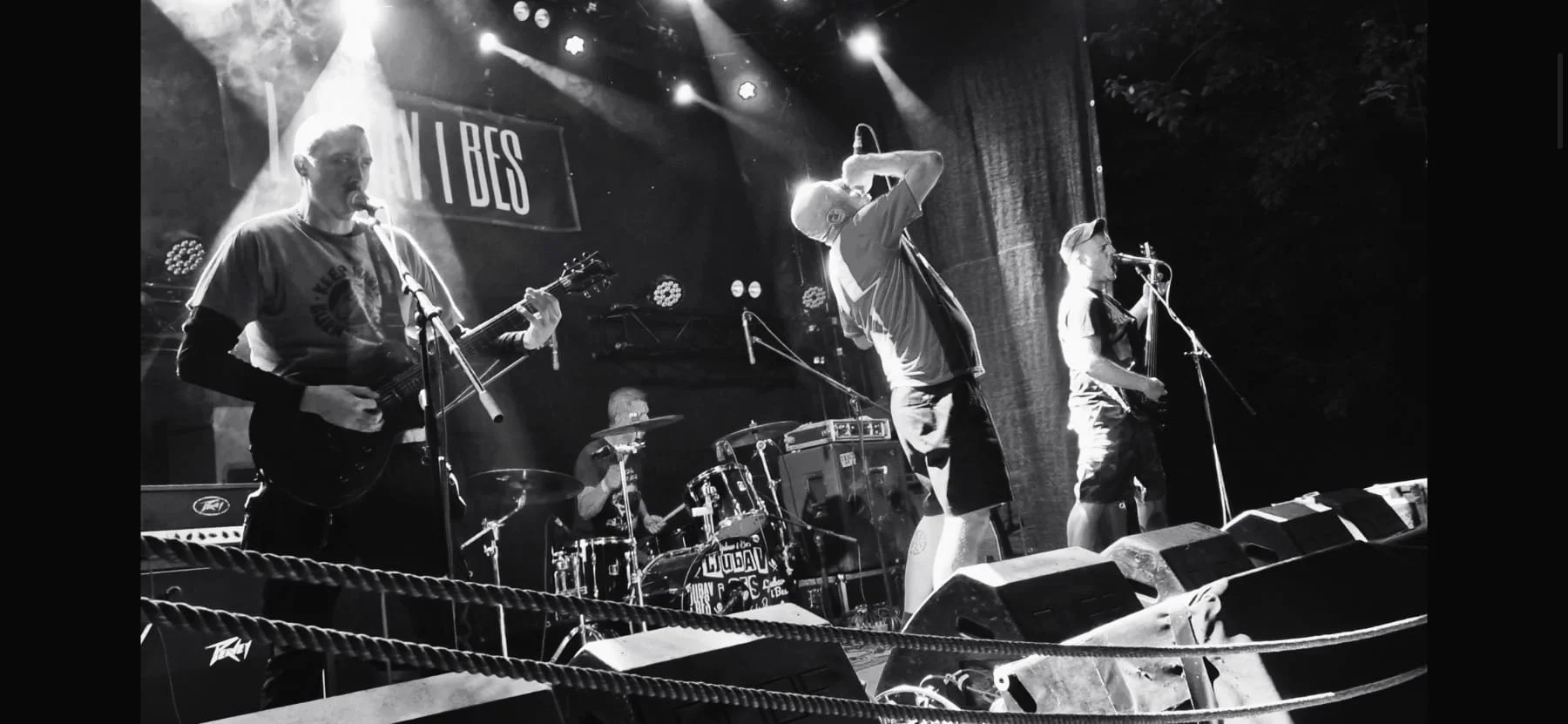 A black and white photo of a band performing on stage, with four members visible: a guitarist singing into a microphone, a drummer, a vocalist holding a microphone to their mouth, and a female bassist. Stage lights illuminate the scene.