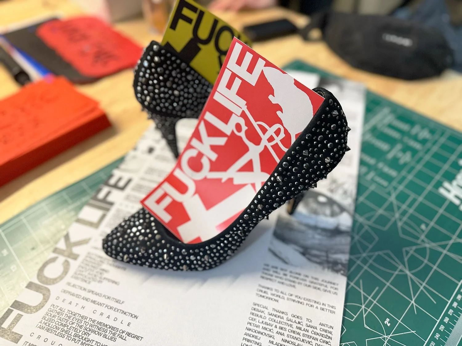 A black high-heeled shoe with silver spikes, holding two cards with bold red and yellow text.