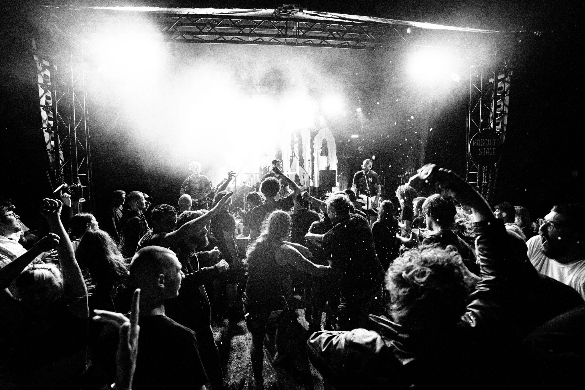 A black and white photo of a live concert with a crowd dancing, musicians on stage under bright lights, and a sign that says 'Mosquito Stage'.