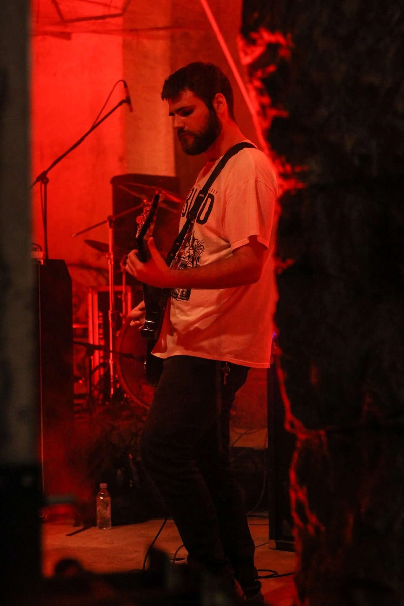 A young man with a beard playing an electric guitar on stage, illuminated by red stage lights, with a microphone stand and sound equipment in the background.