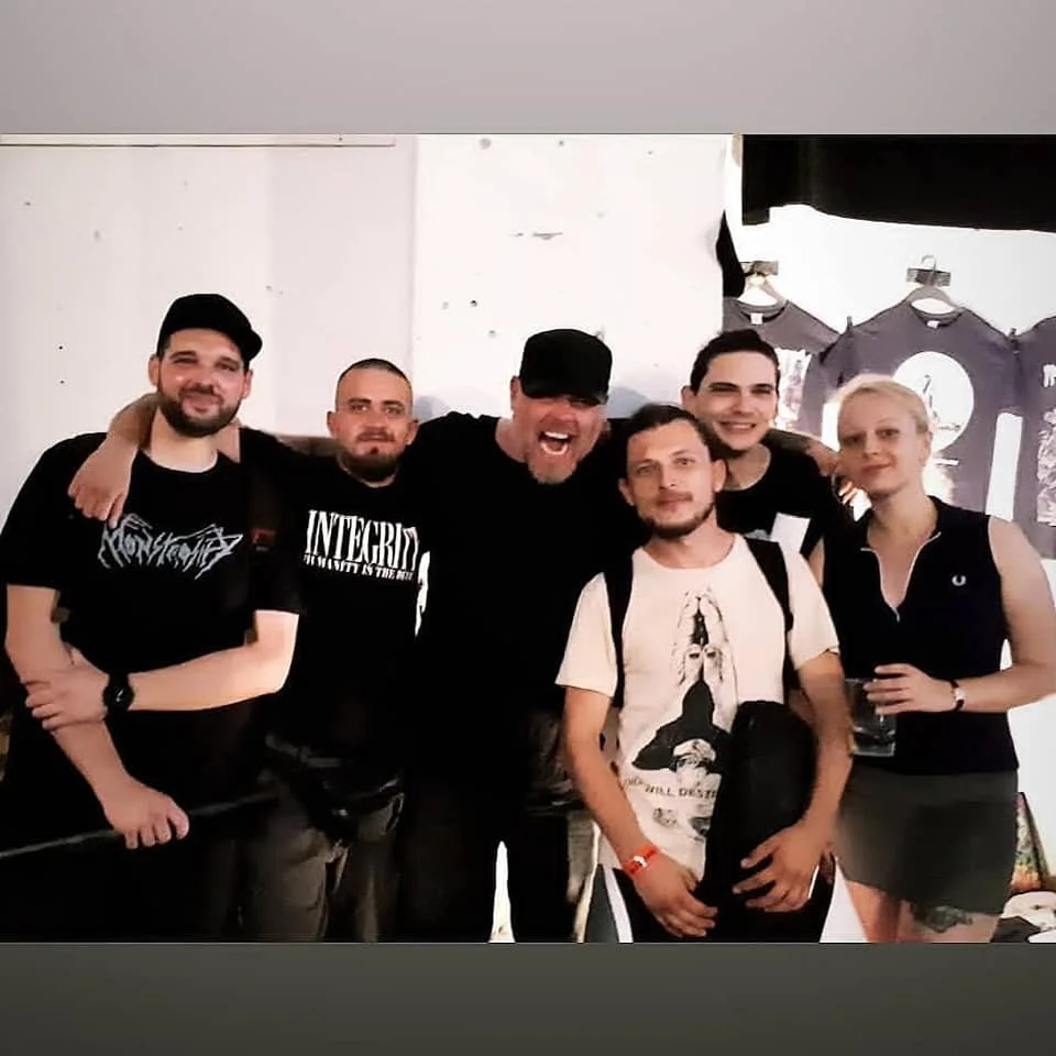 Group of six people smiling and posing together indoors, some with drinks in hand, with t-shirts and casual attire.