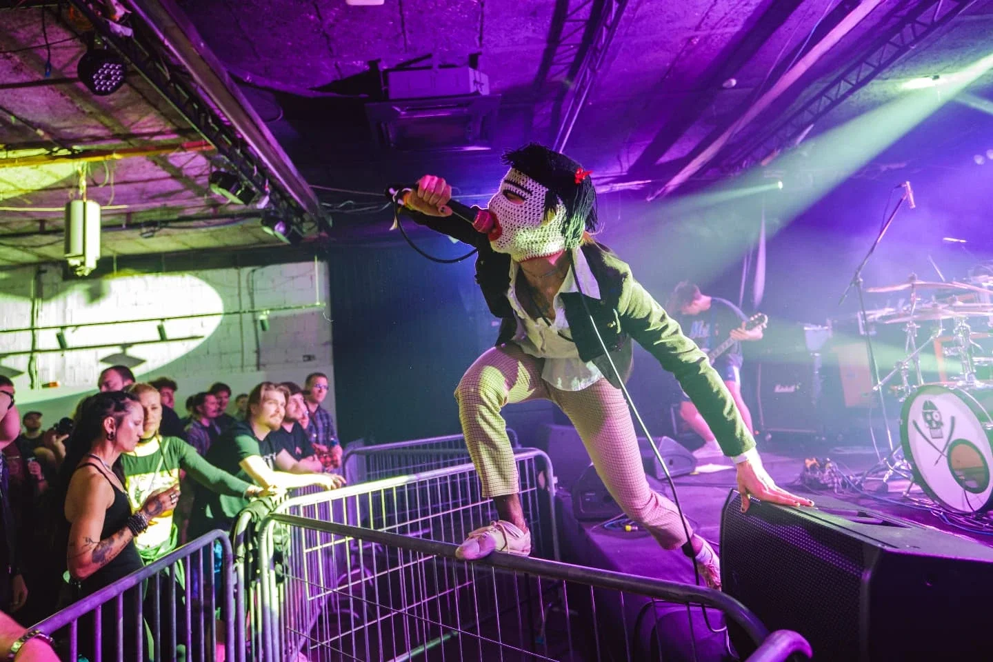 Singer wearing a mask on stage, kneeling with one hand on the stage monitor, performing at a concert with an audience watching.