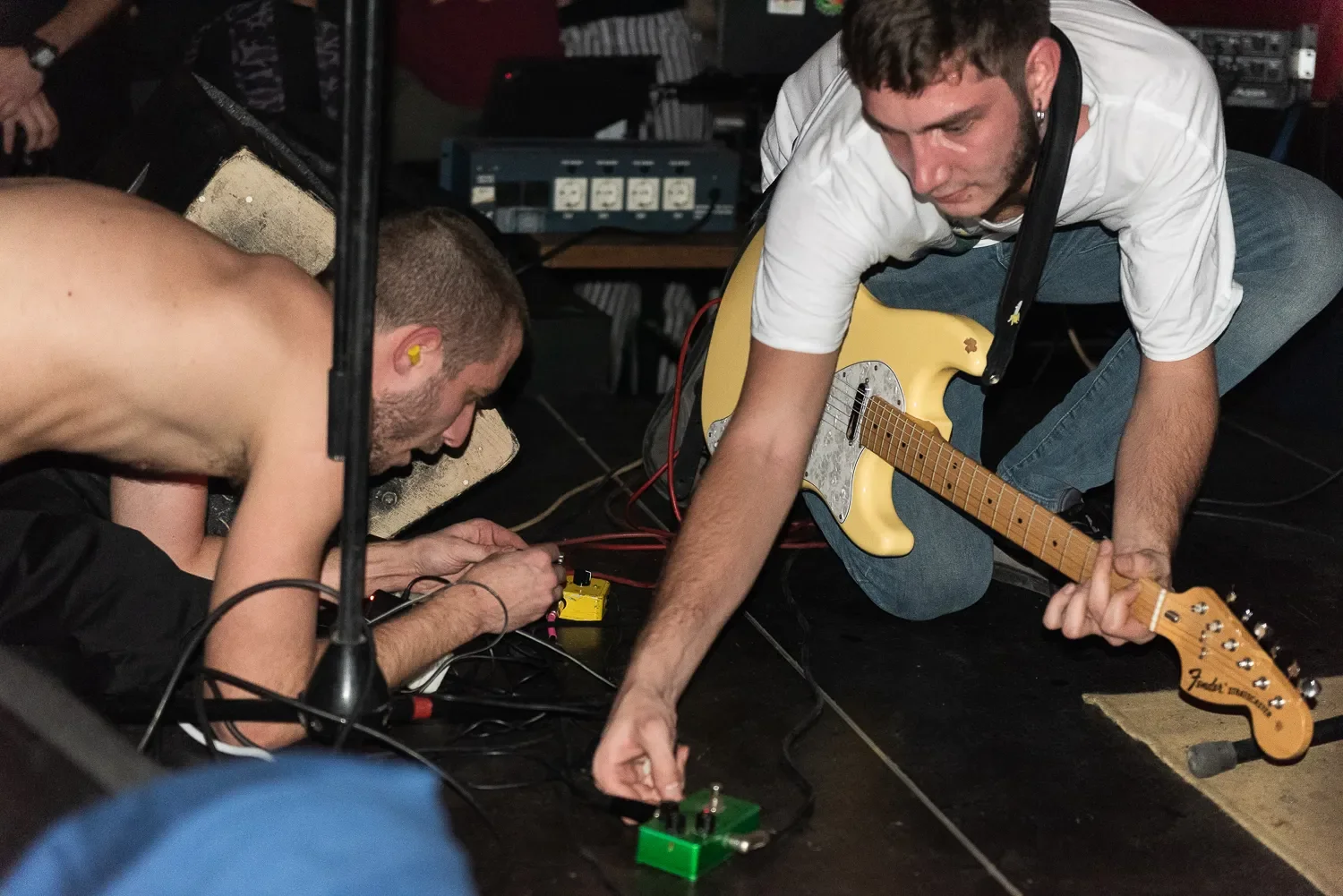 Two men are on the floor working with music equipment. One is shirtless and the other is wearing a white t-shirt and jeans, holding a yellow Fender electric guitar. They are connecting or adjusting audio gear surrounded by cables and electronic devic