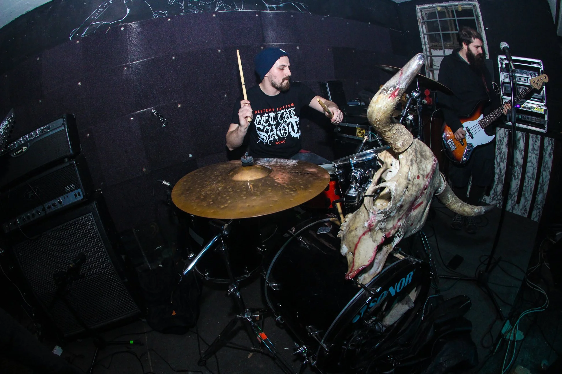 Two musicians performing on stage; one is playing drums and the other is playing bass guitar. There is a large animal skull with horns as stage decoration in front of the drum kit.