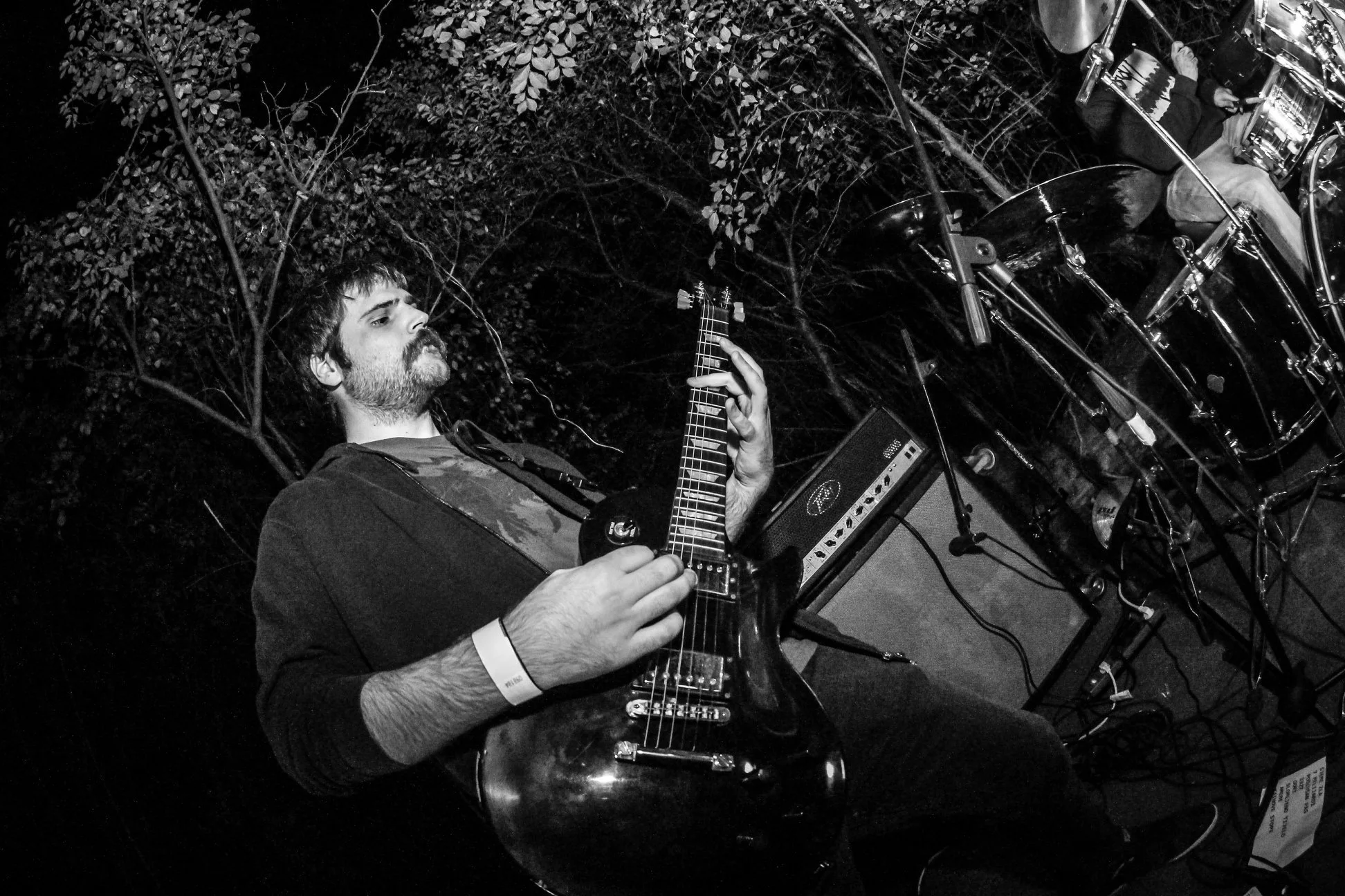 A man playing an electric guitar on stage at night, with a drum set and music equipment behind him, under trees.