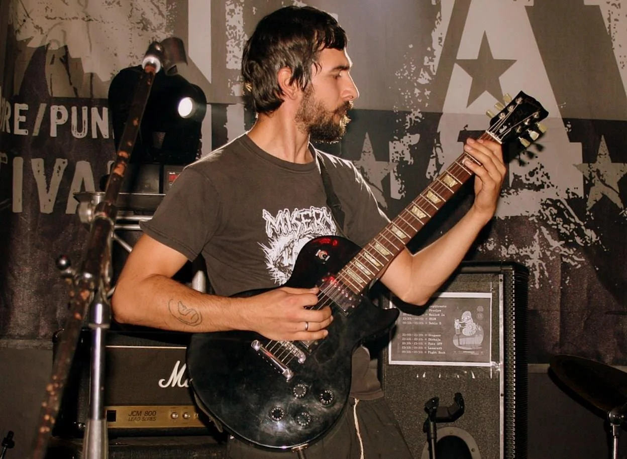 A man with dark hair and a beard playing a black electric guitar on stage at a music event.
