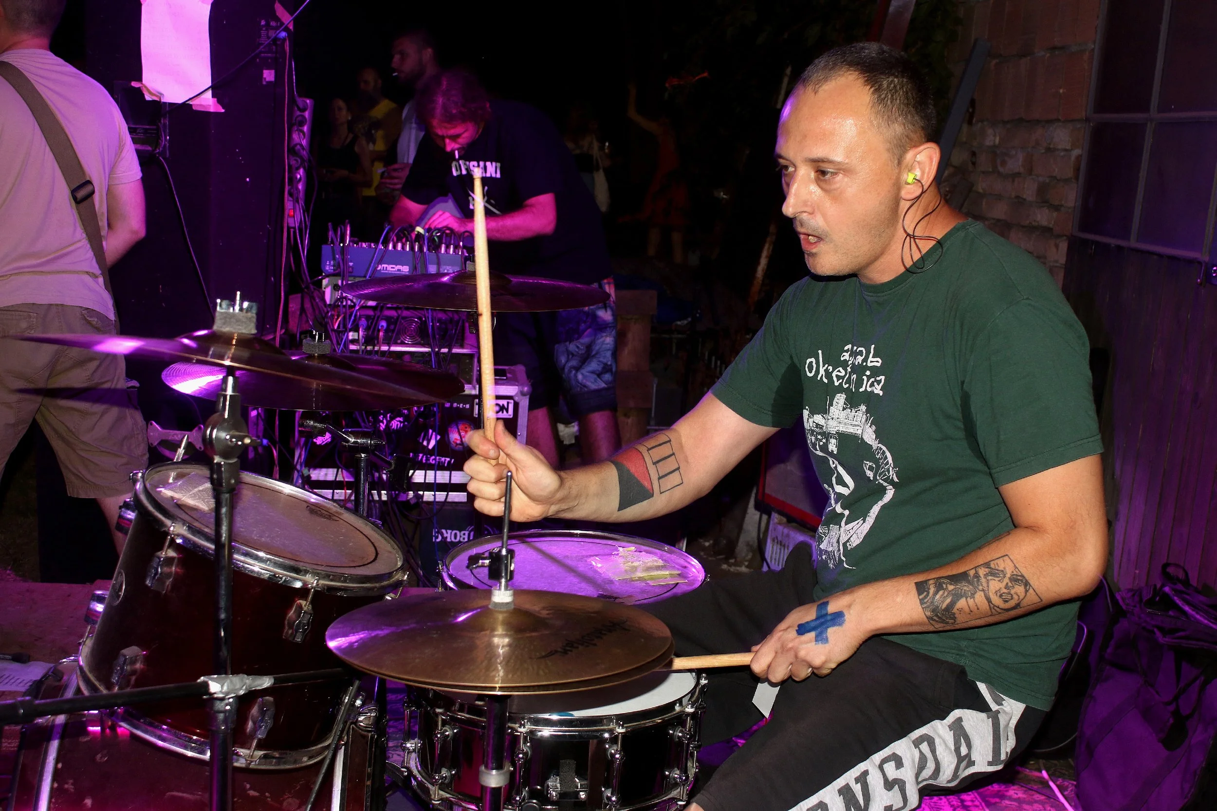 A musician playing drums at an outdoor night event, with equipment and other people in the background.
