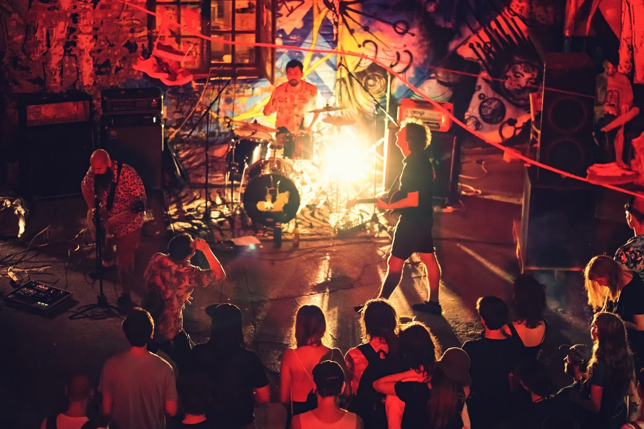 A live music performance with a band playing on stage, illuminated by warm lighting, featuring a drummer, a guitarist, and an audience watching in a dimly lit indoor venue with graffiti-style artwork on the walls.