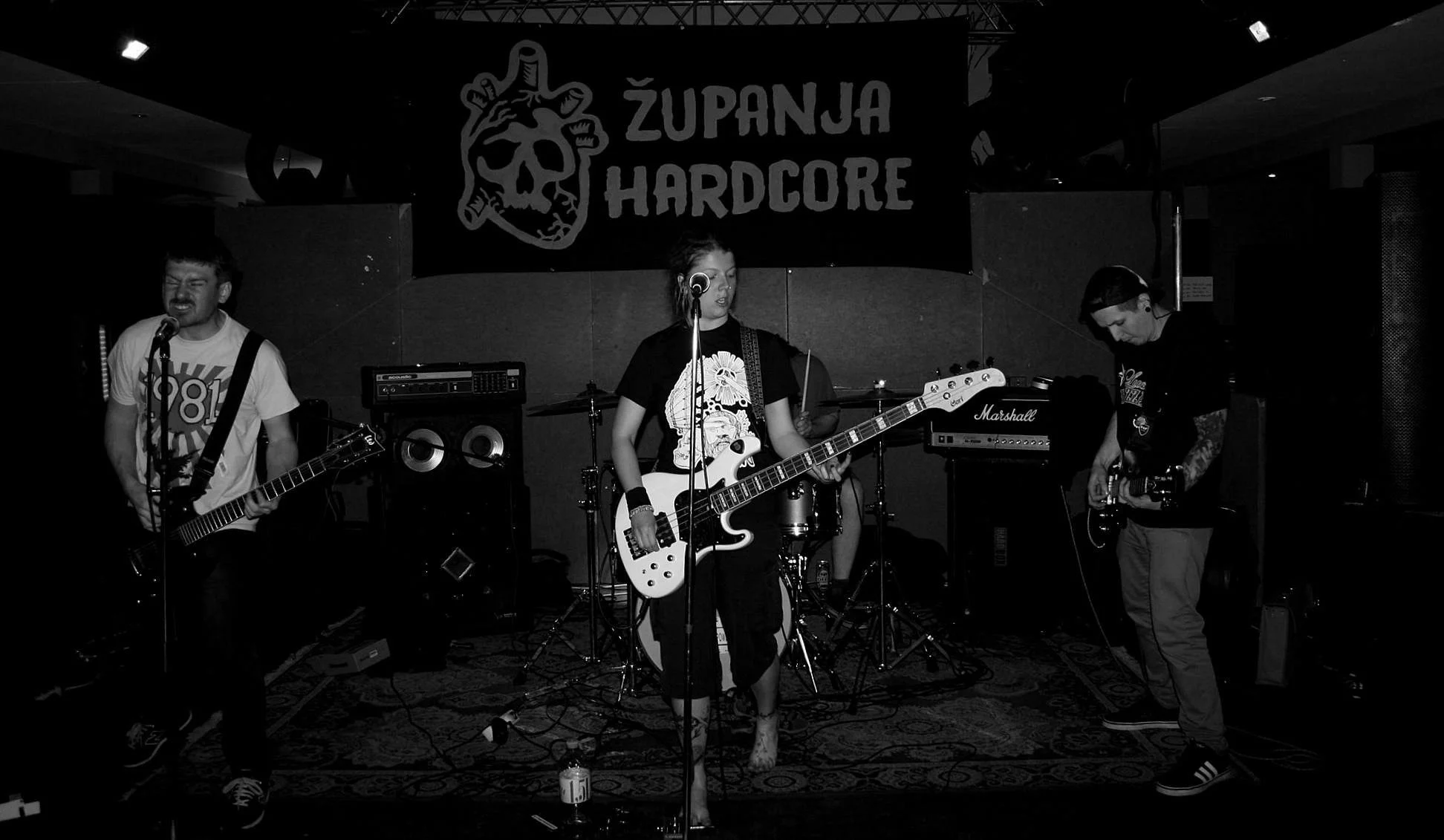 A black and white photograph of a band performing on a stage with a banner that says "Županija Hardcore" in the background. There are three musicians visible, one playing the guitar on the left, a female bassist in the middle, and another guitarist o
