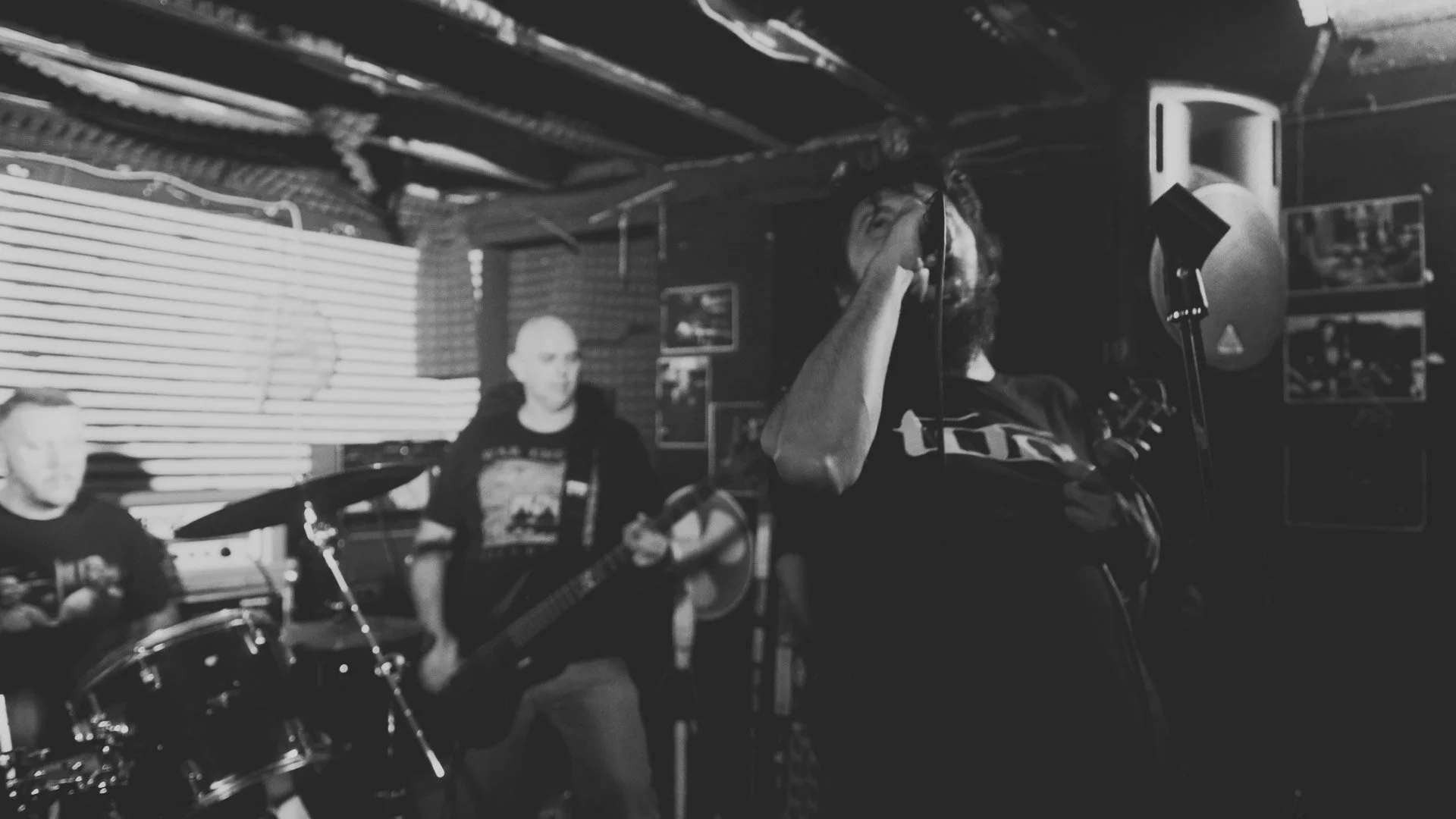 A black and white photo of a band performing in a dimly lit indoor venue. The image shows a drummer on the left, a person with a guitar in the center, and another person with a microphone on the right.