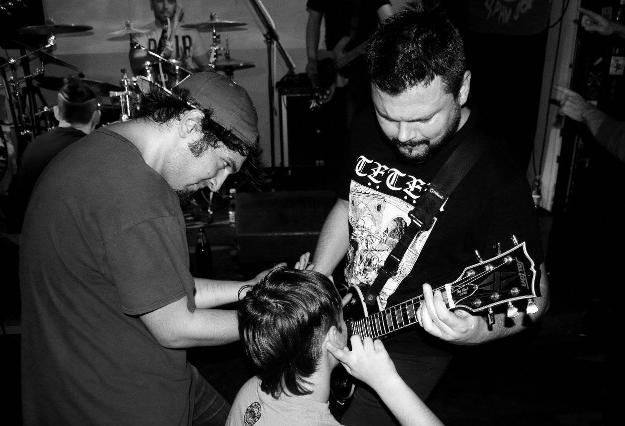 Black and white photo of a man playing an electric guitar, a child touching his face, and another person leaning in close, with a band playing in the background.
