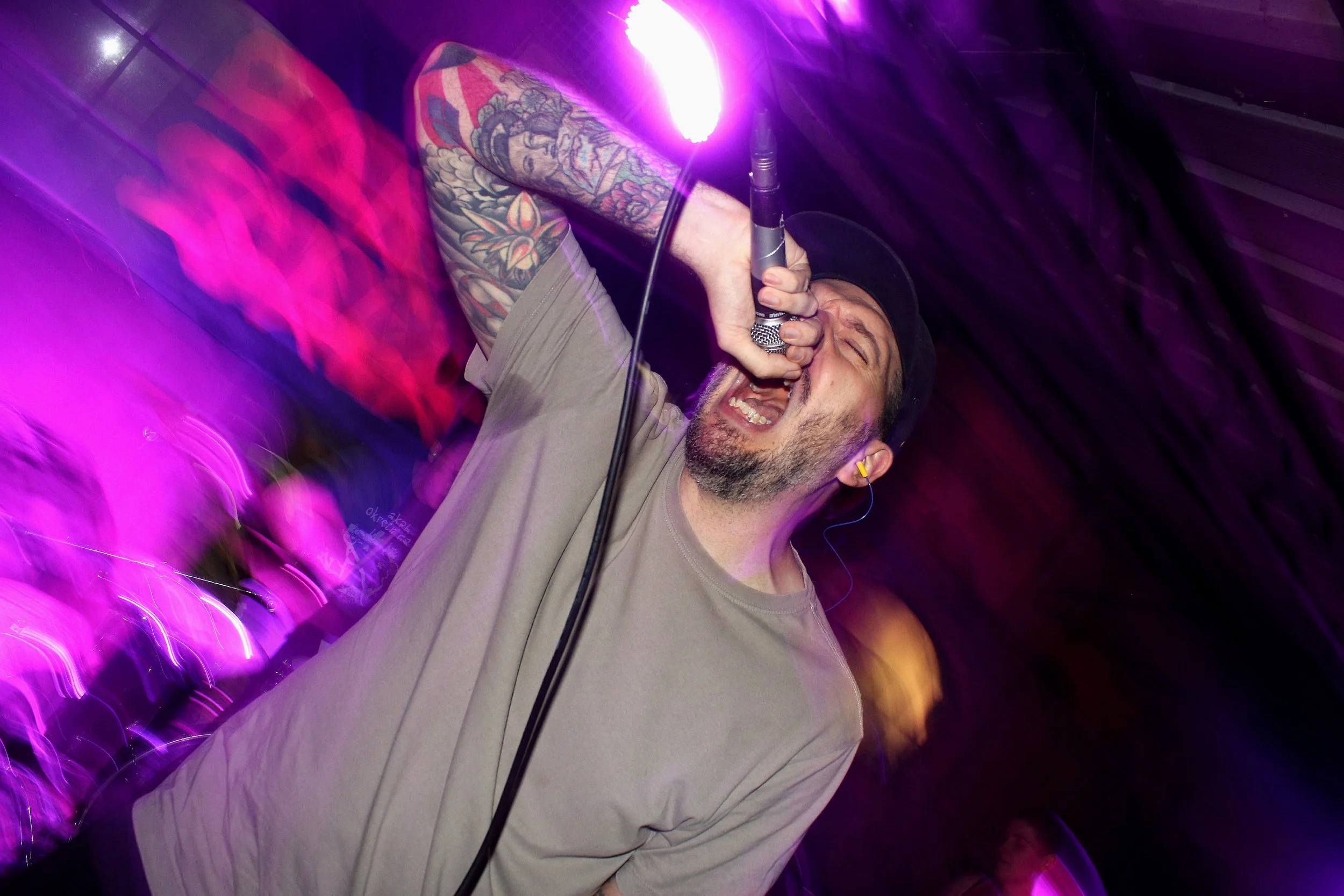 A man singing into a microphone with a big smile, wearing a black cap and gray t-shirt, with tattooed arms, under colorful pink and purple stage lights.