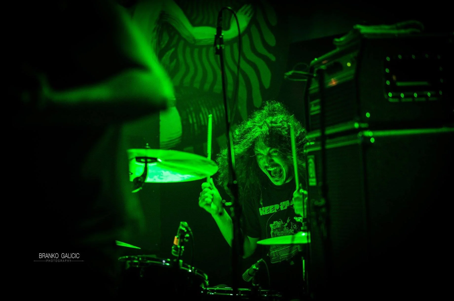 A person with long curly hair passionately singing or screaming into a microphone while playing drums in a dark room illuminated with green light.