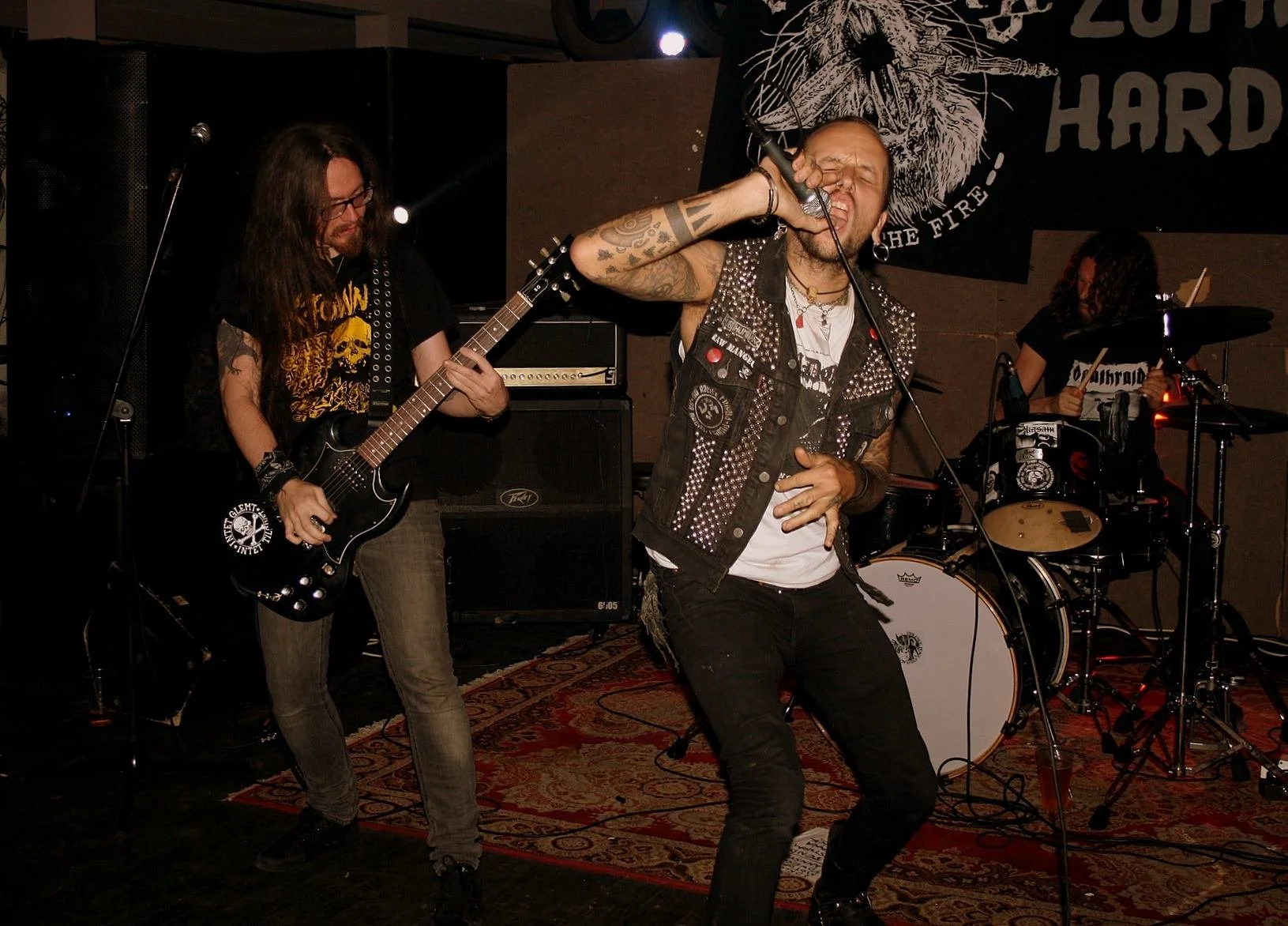 A band performing live, featuring a vocalist passionately singing into a microphone, a guitarist playing an electric guitar, and a drummer playing on a drum kit, all on stage at a music venue. Band name: Crutches