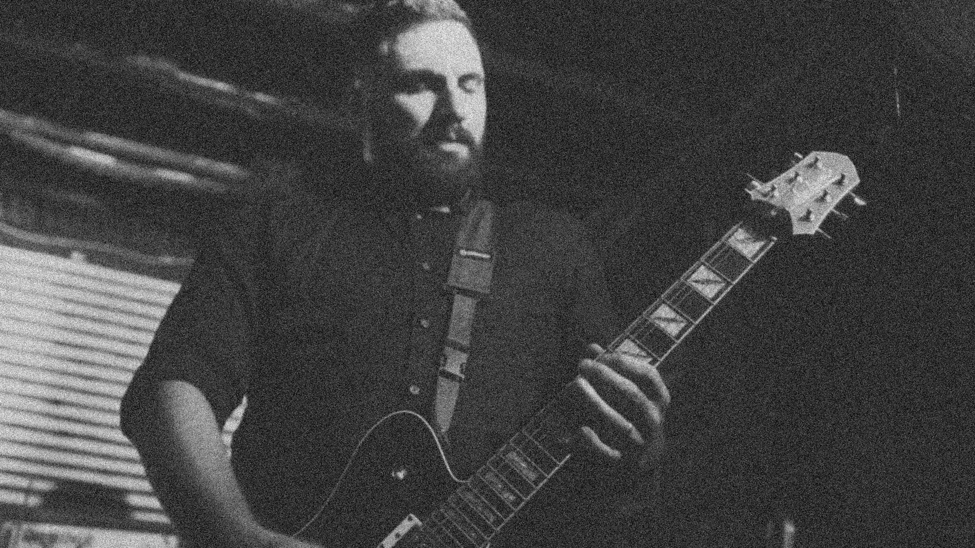 A person with long hair and a beard playing an acoustic guitar in a dimly lit room.